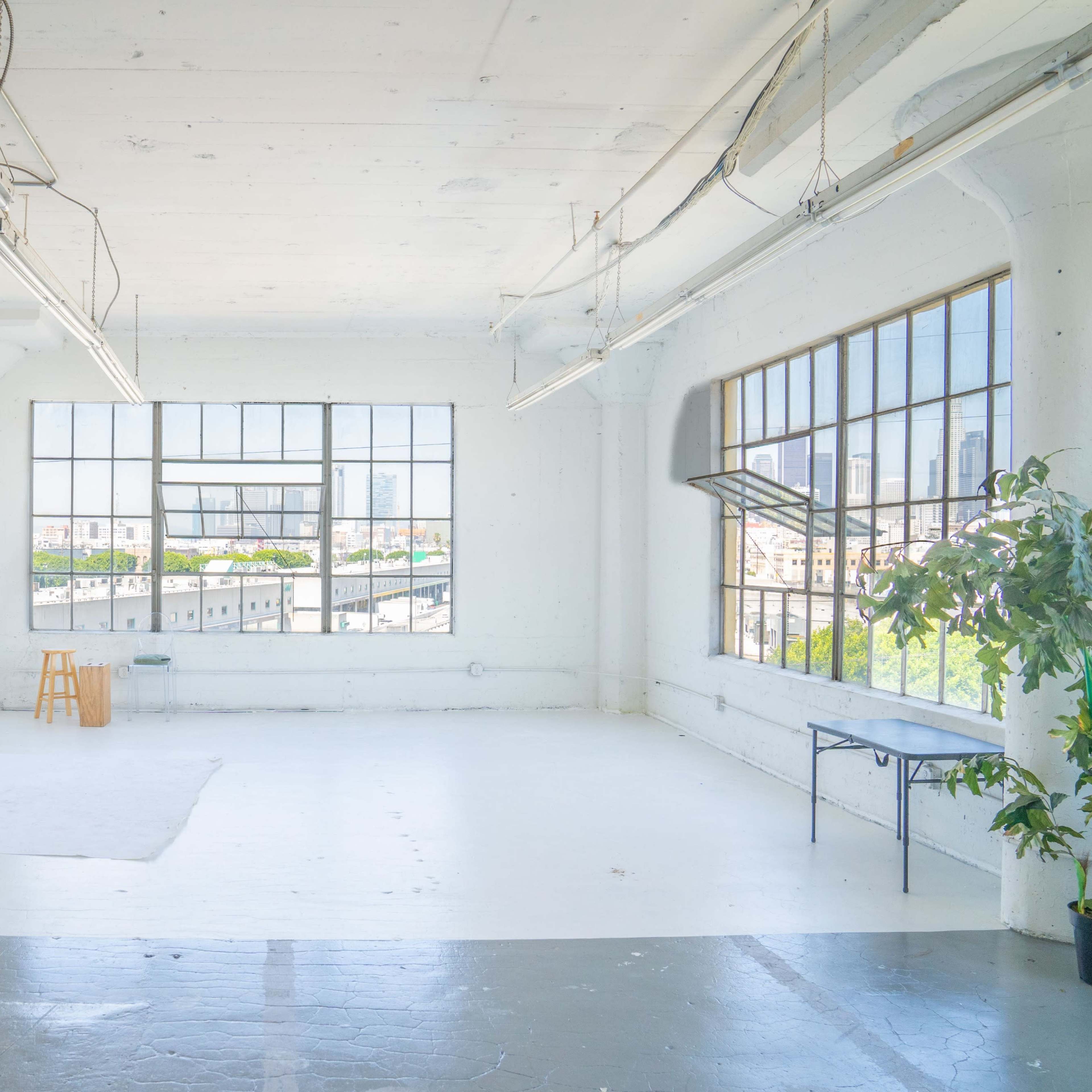 A spacious, well-lit studio with large windows, a white backdrop on a stand, a stool, and a table against the walls.