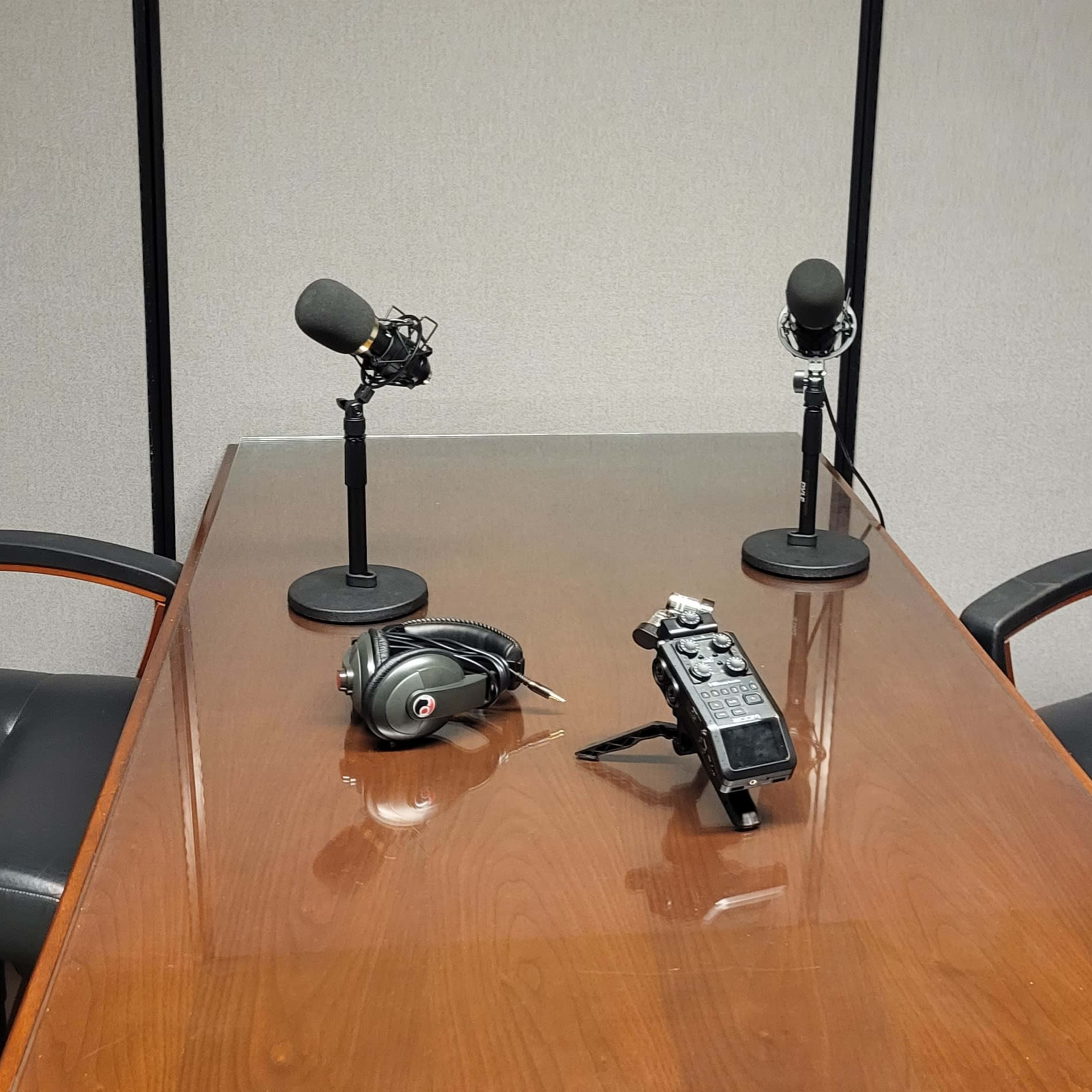 A conference room is set up with two microphones on stands, a recording device, and two black chairs around a wooden table.