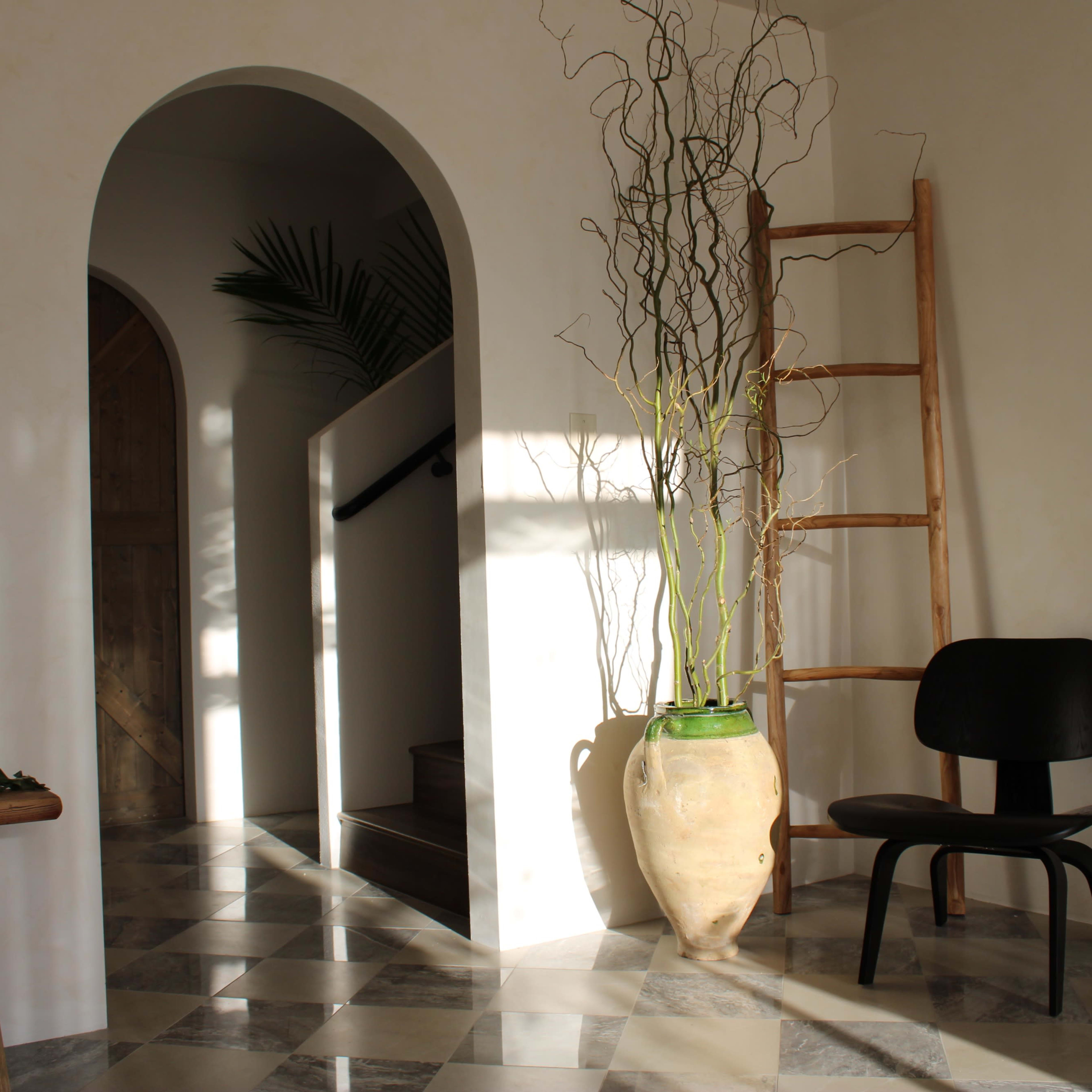 A minimalist interior with a wooden ladder, a black chair, a large vase with branches, and a staircase leading to a doorway.