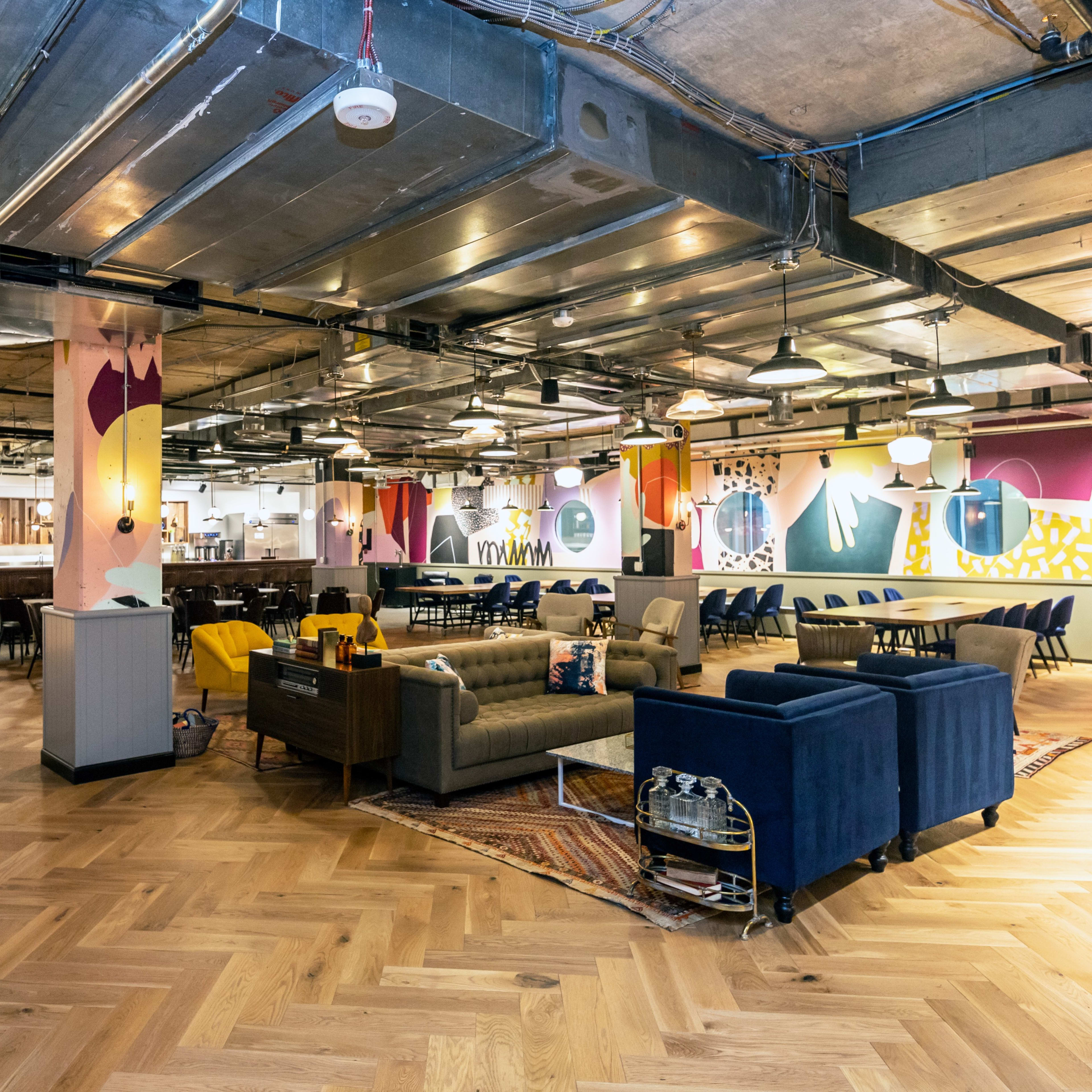 A spacious, modern interior of a communal workspace with wooden flooring, various seating areas, and colorful wall art.