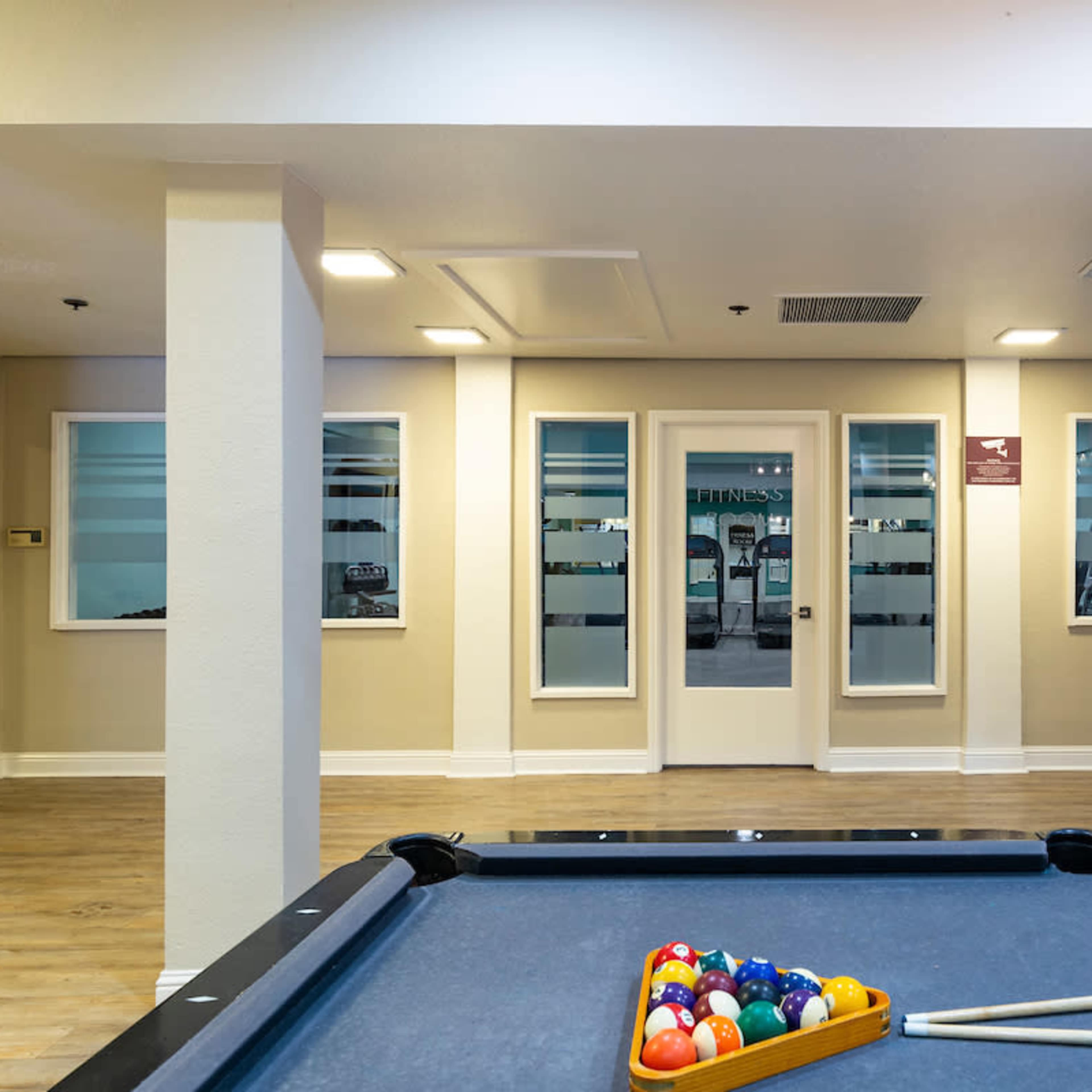 A billiard table with a triangle of colored balls is in the foreground, while a fitness center door is visible in the background.