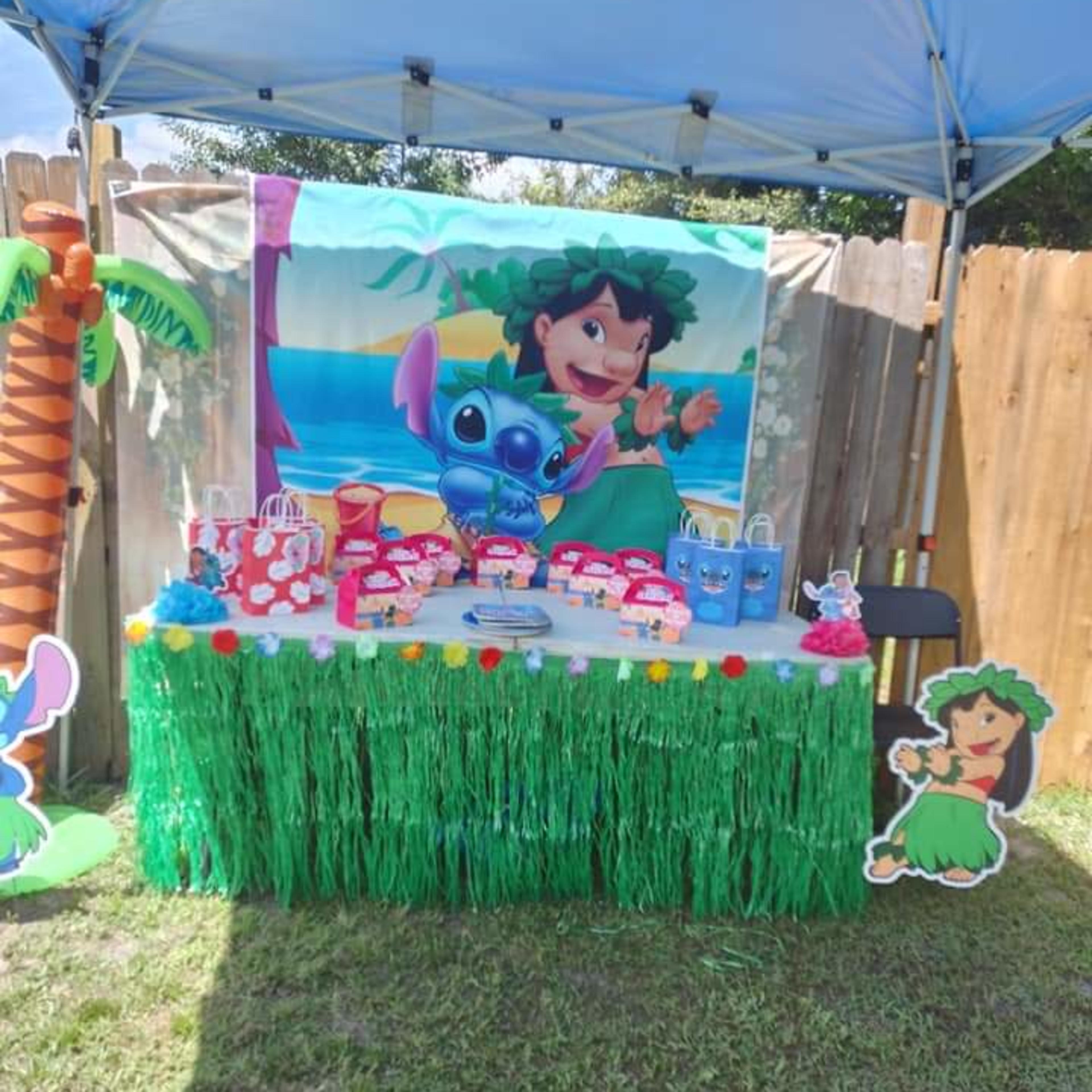 A decorated table with a green grass skirt, featuring themed party supplies and a backdrop of a cartoon character, sits under a blue canopy in a grassy area.