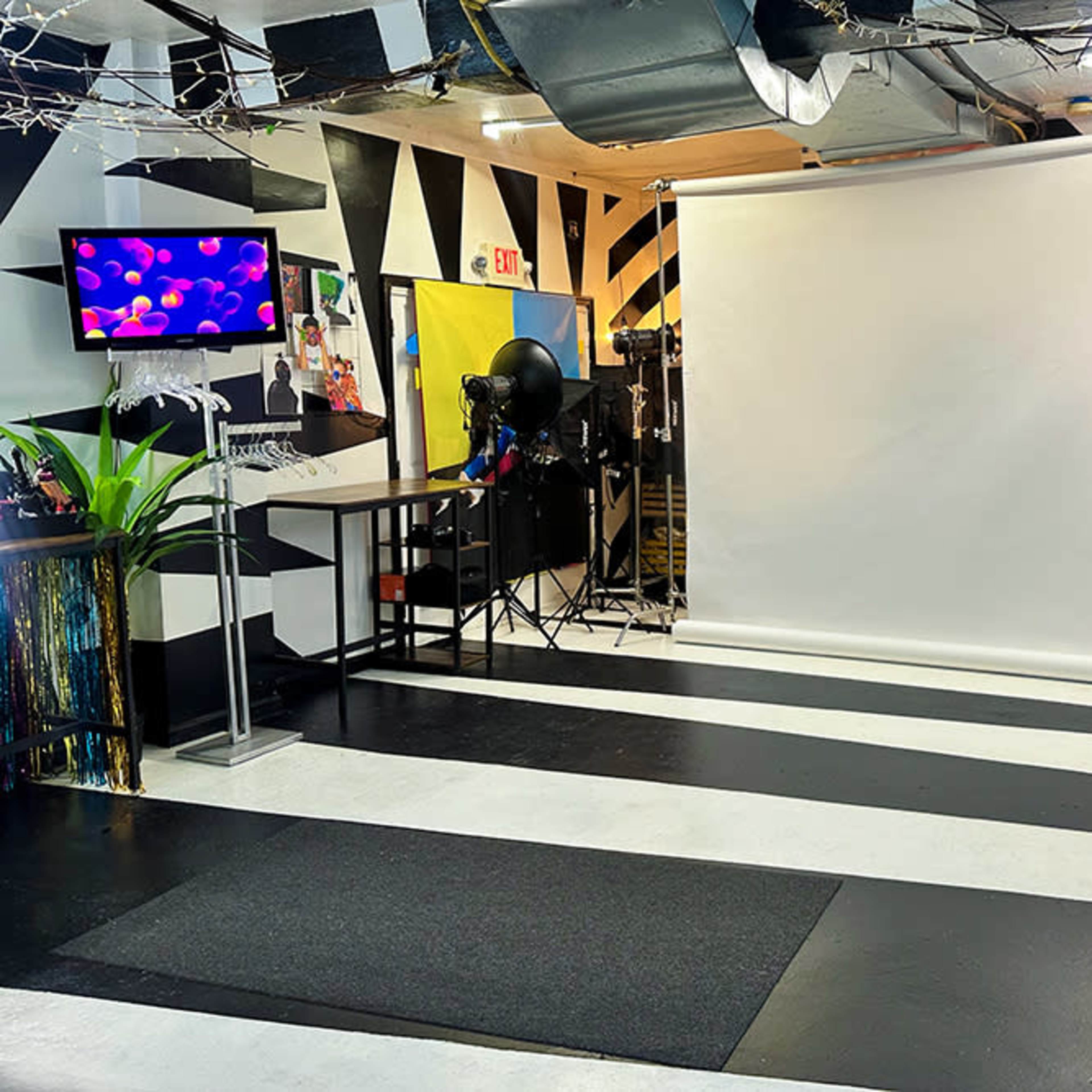 A photography studio with a blank backdrop, striped flooring, and various props and decorations in the background.