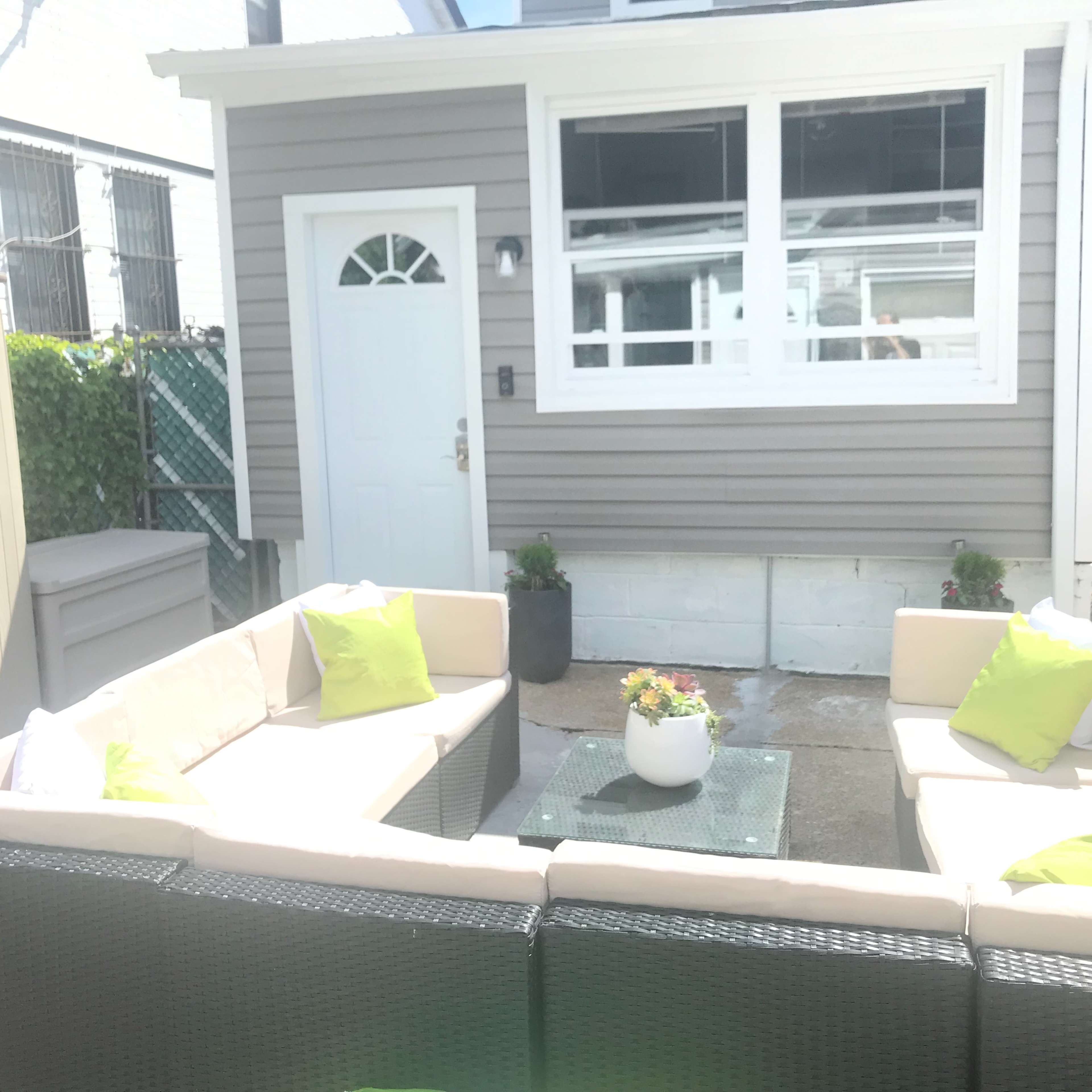 A small outdoor seating area with two light-colored sofas arranged around a glass coffee table, situated in a courtyard beside a house.
