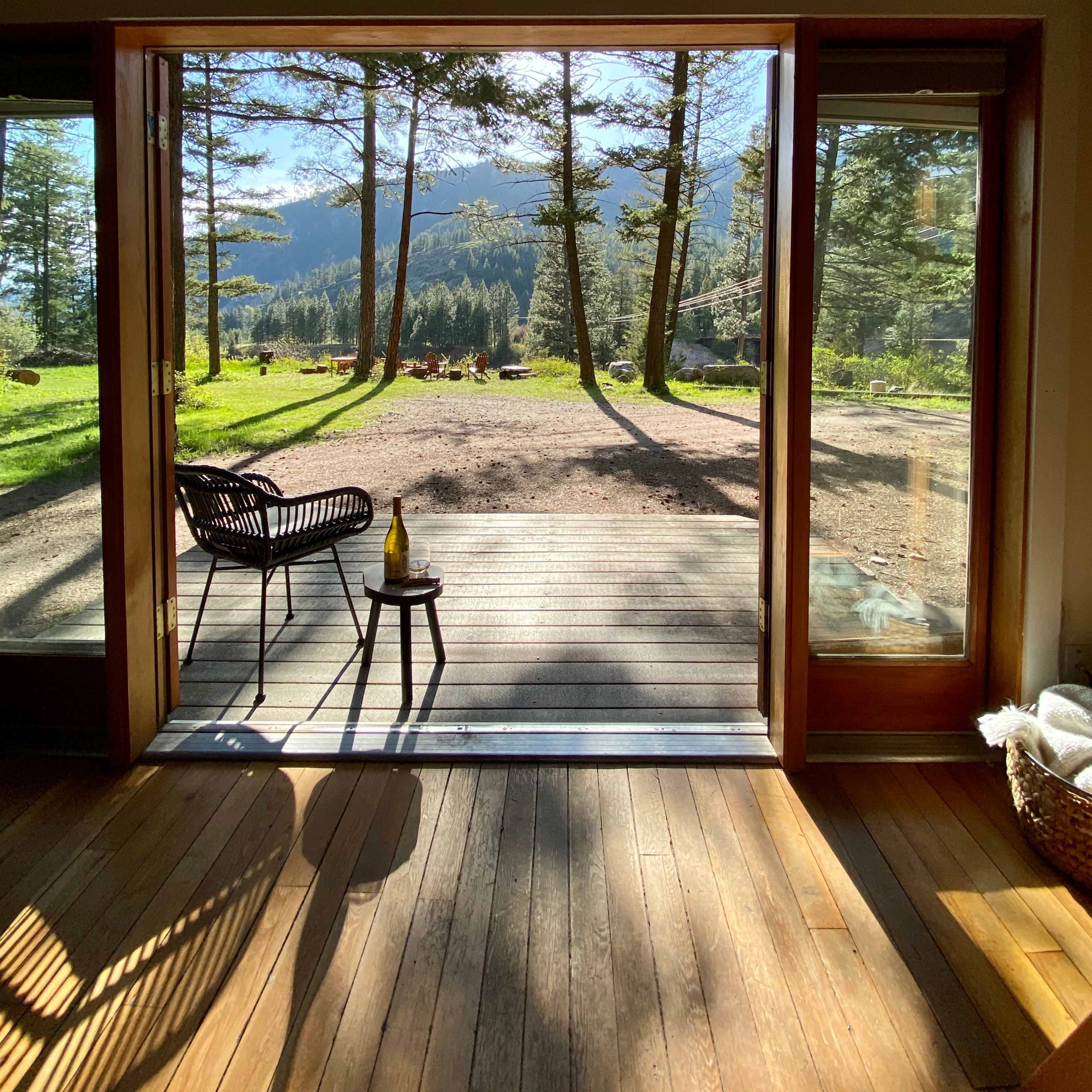 A wooden deck extends from an open doorway, framed by trees and mountains in the background.