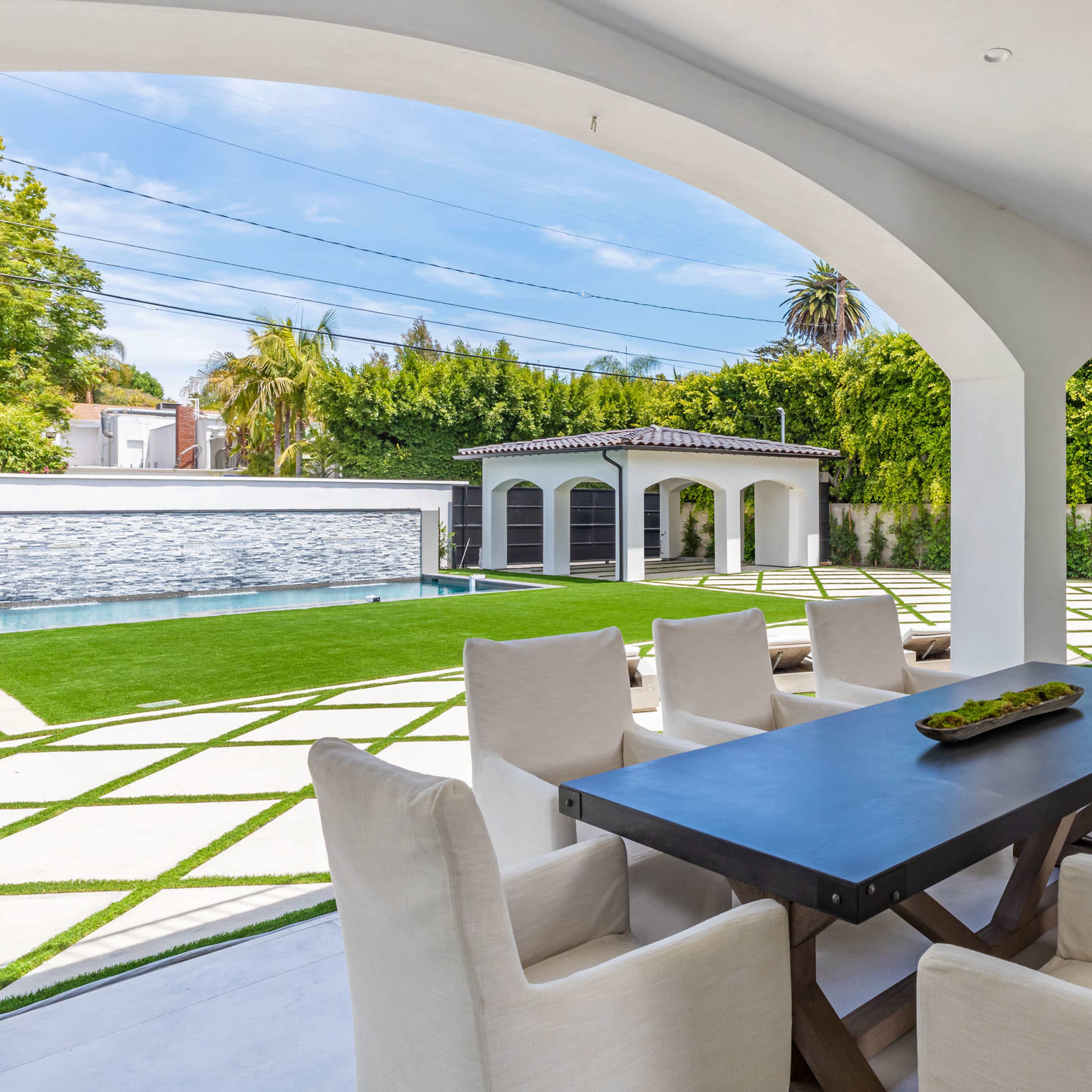 A spacious outdoor patio area with a large dining table surrounded by white chairs, overlooking a landscaped yard with a pool and a separate structure in the background.