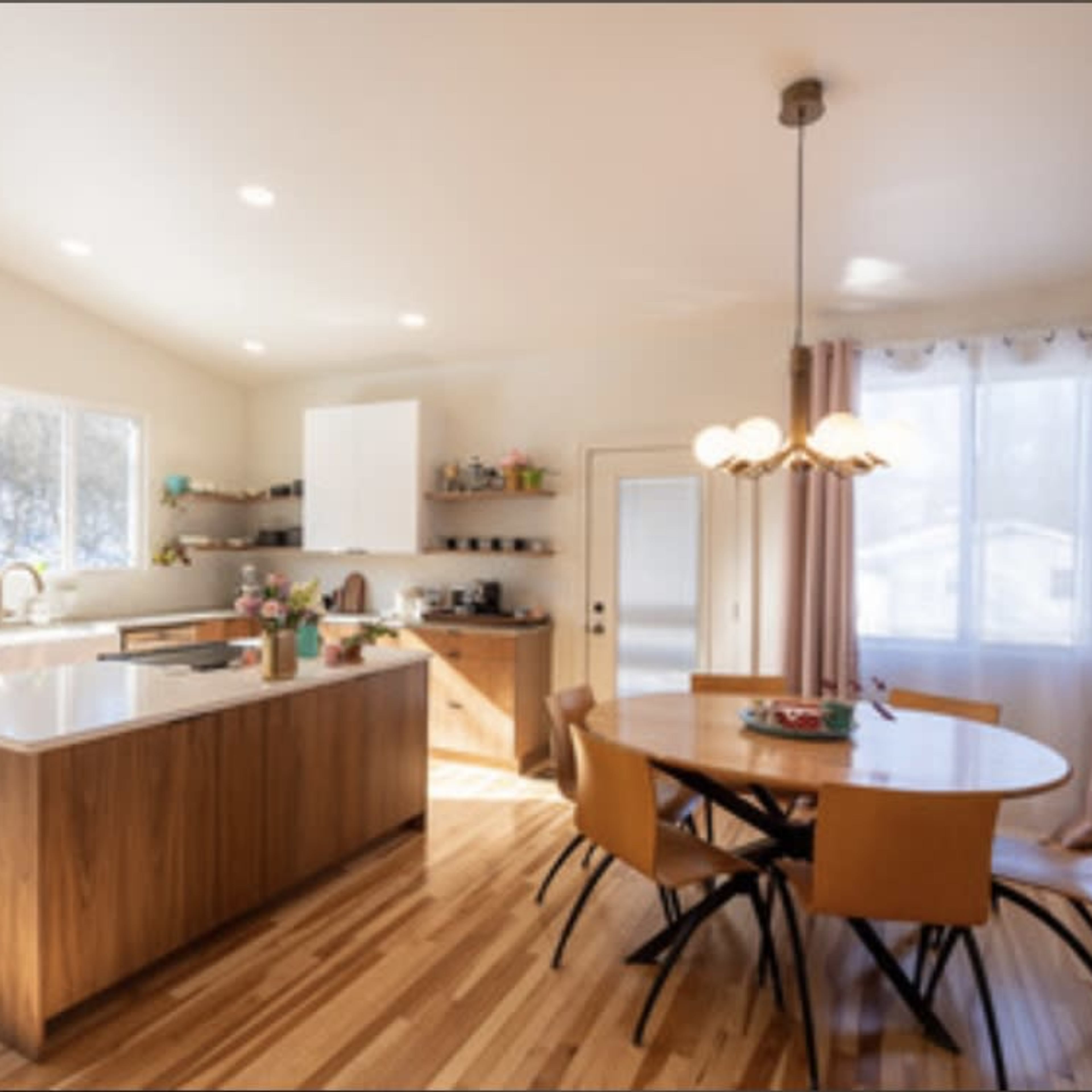 A modern kitchen features an island with a sink, wooden cabinetry, and a dining table surrounded by chairs, illuminated by natural light from large windows.