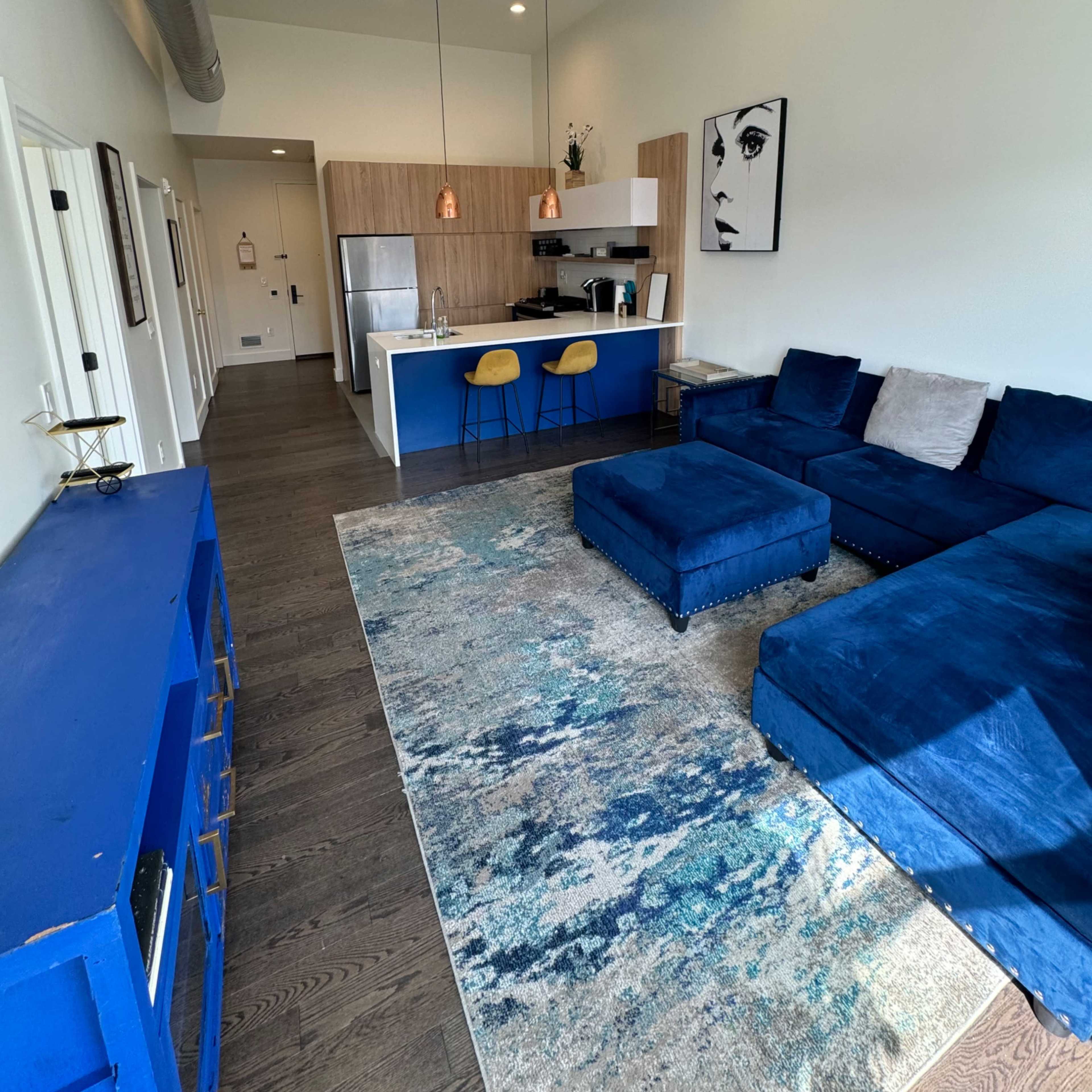 A modern living room featuring a blue sectional sofa, a matching ottoman, a large area rug, and a kitchen area with wooden cabinets and bar stools.