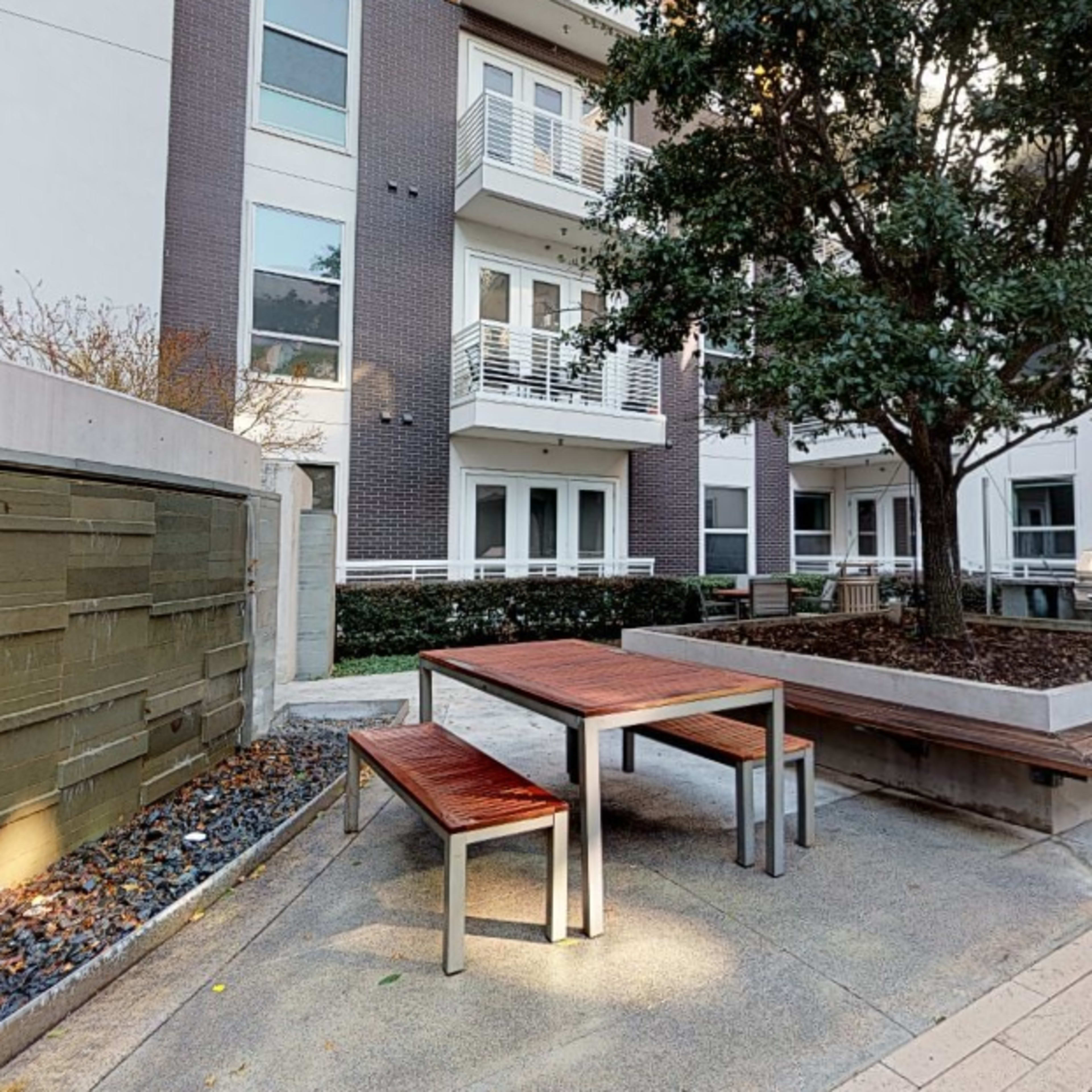 A courtyard area with a large tree, wooden tables, and benches surrounded by modern apartment buildings.
