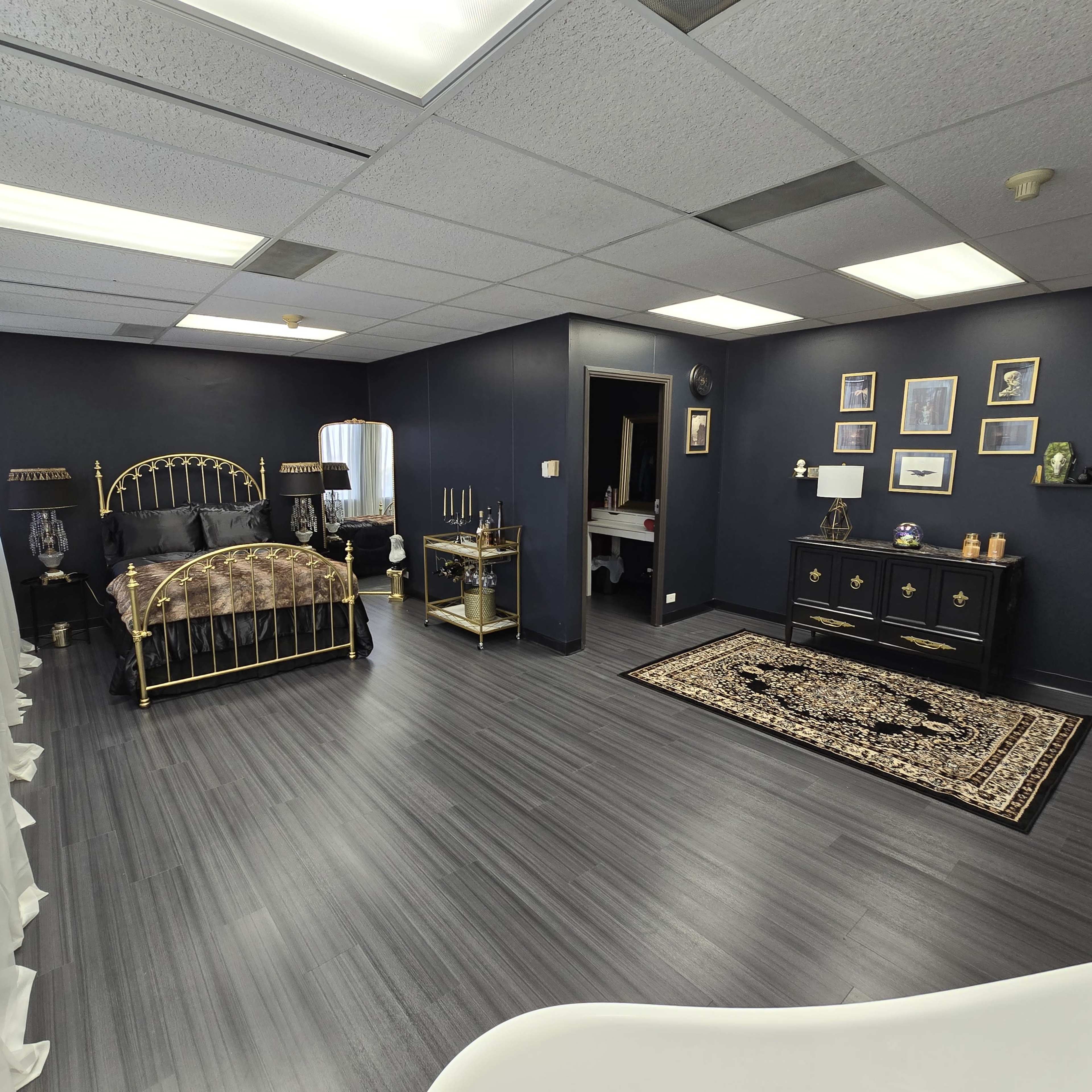 A spacious bedroom featuring a gold bed frame, dark blue walls, a black dresser, and a decorative area rug.