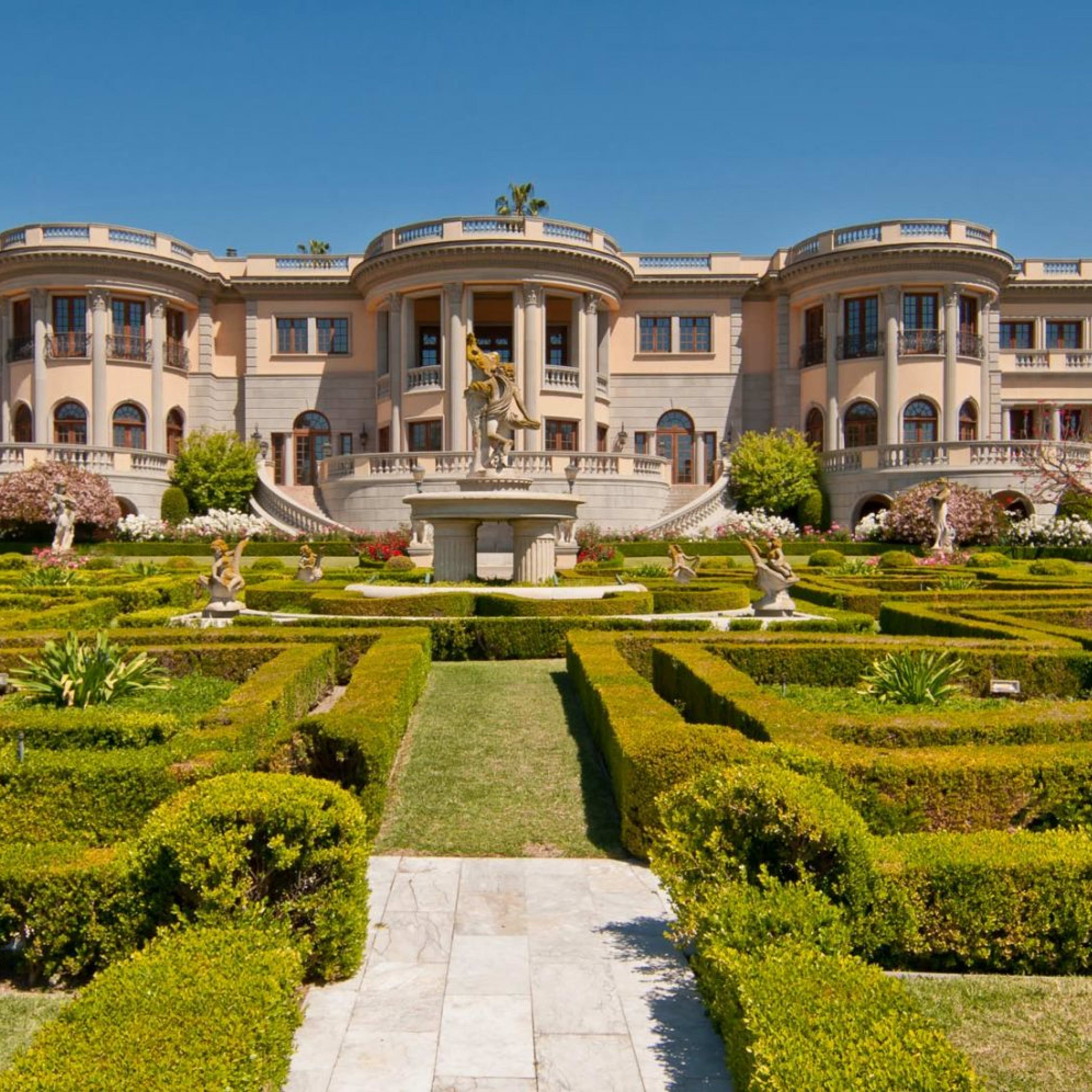 A large, elegant mansion is situated behind a meticulously designed garden featuring neatly trimmed hedges and decorative sculptures.