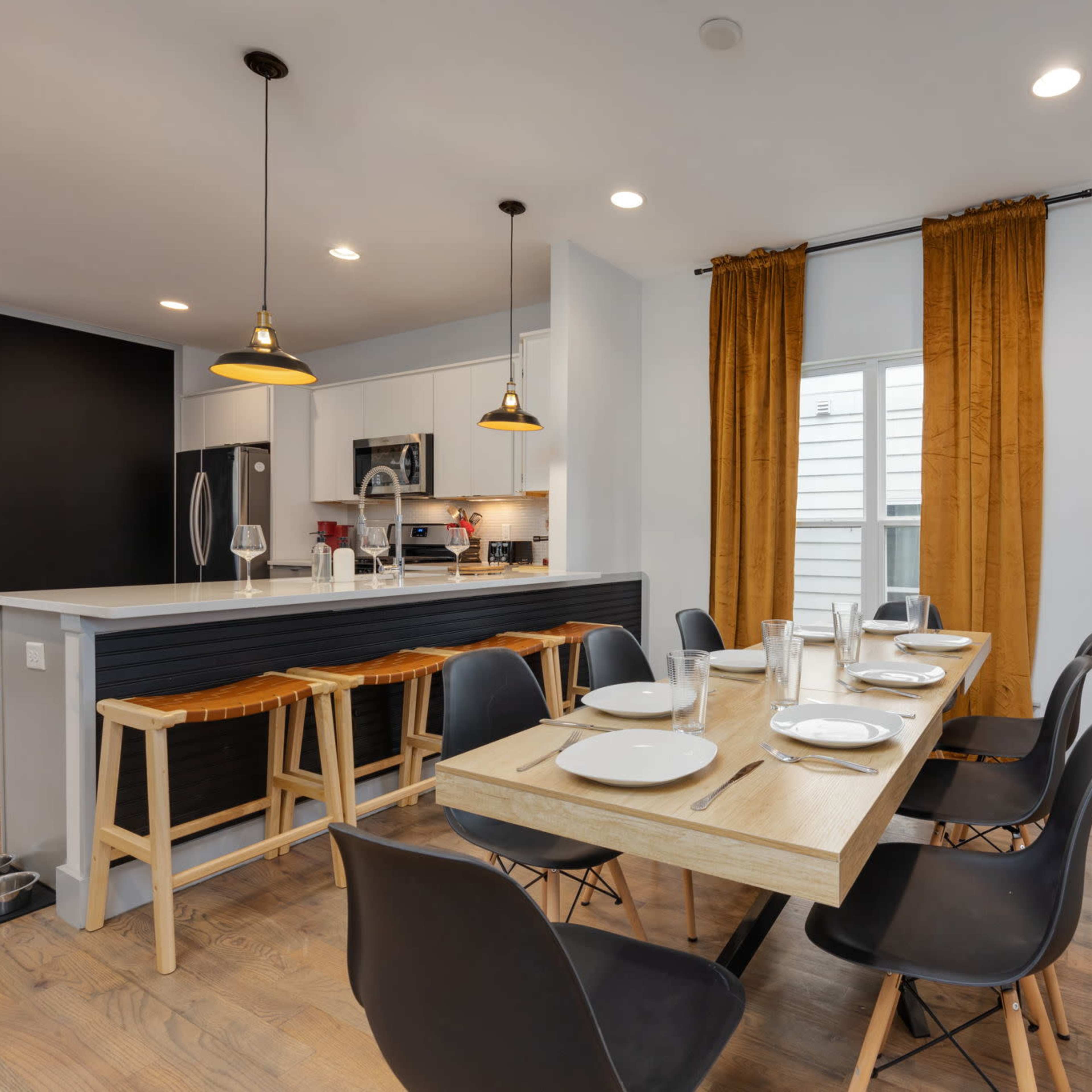 A modern dining area features a wooden table set for six, complemented by black chairs, and a kitchen with white cabinetry and a bar area with stools.