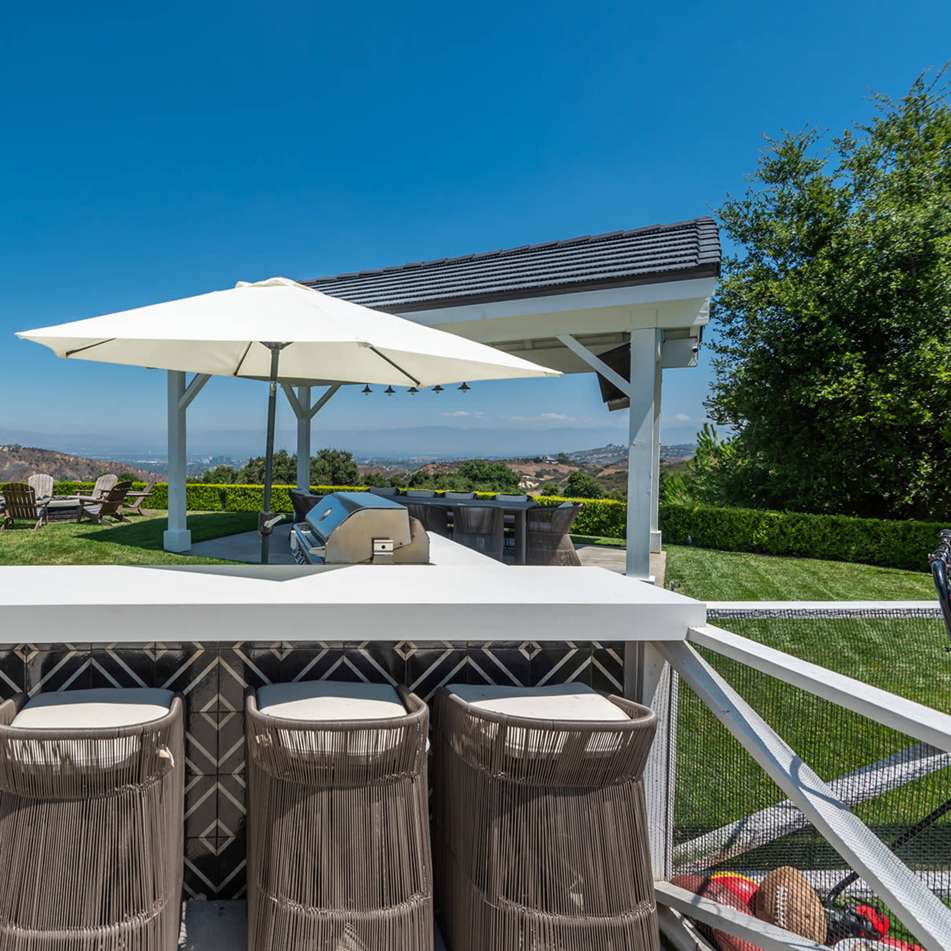 A outdoor bar area with four stools, a grill, and large umbrellas overlooking a green lawn and distant hills.