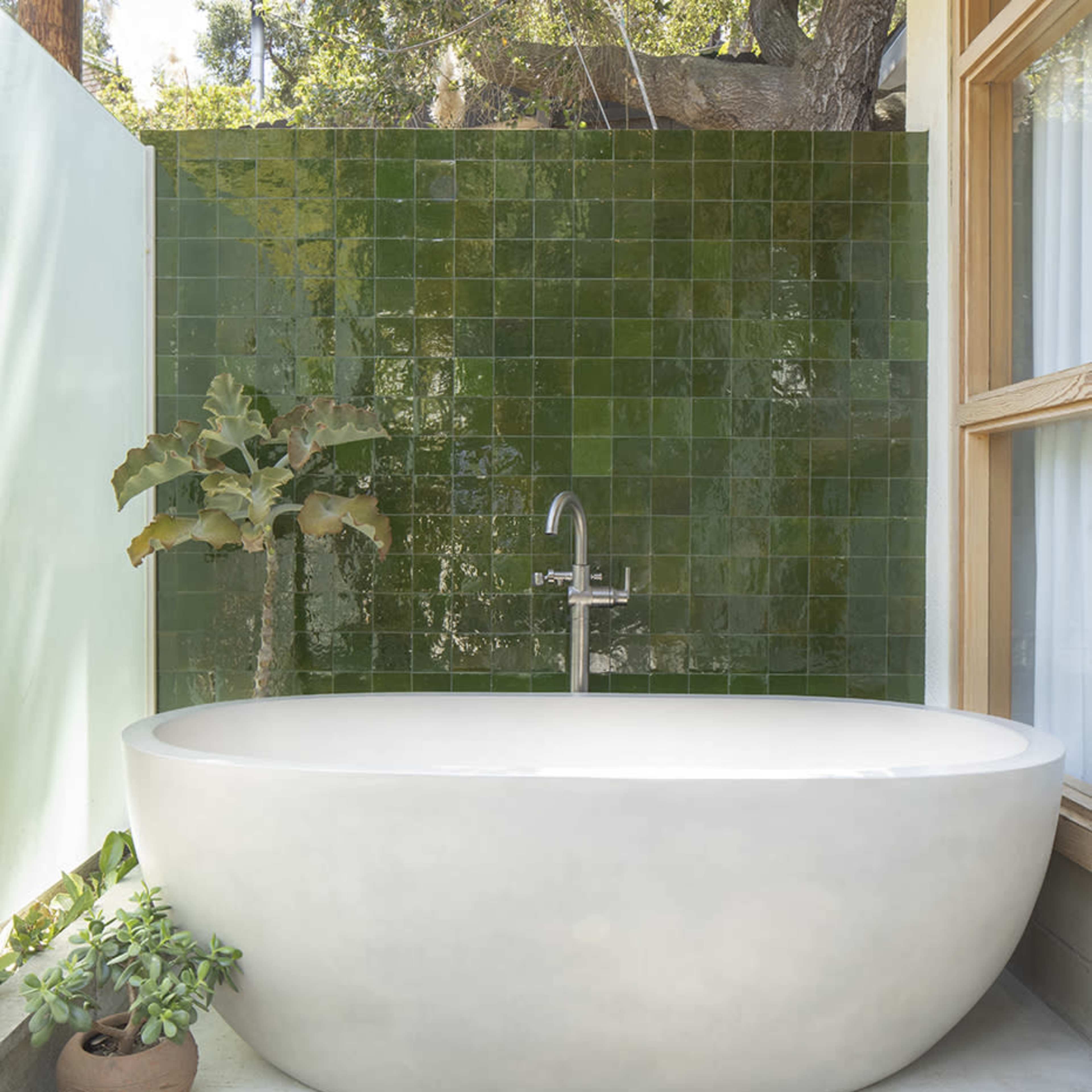 A modern outdoor bathroom featuring a freestanding oval bathtub, a green tile backdrop, and a pebble-stone floor.
