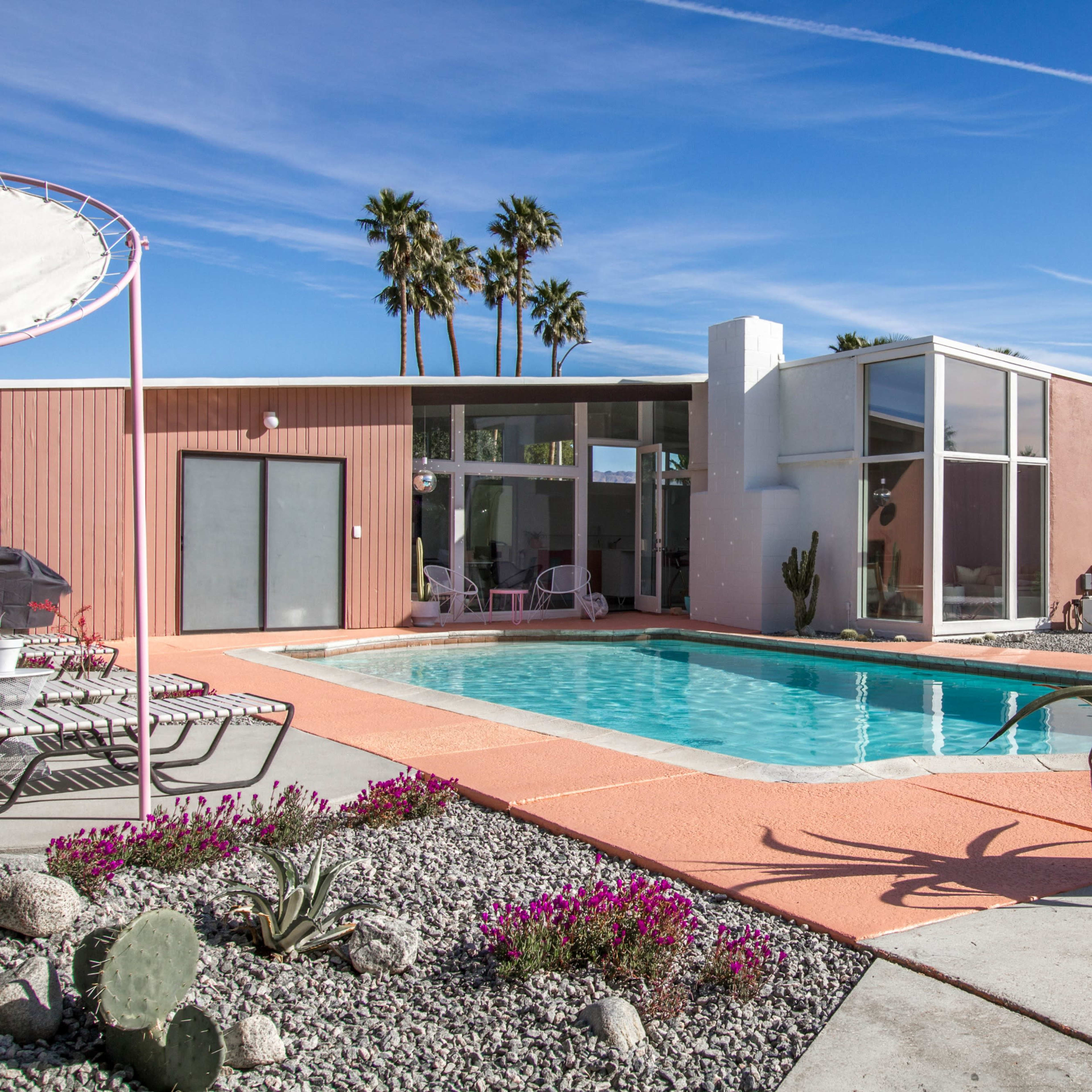 A modern house with large windows surrounds a swimming pool, featuring a pink exterior and desert landscaping with palm trees.