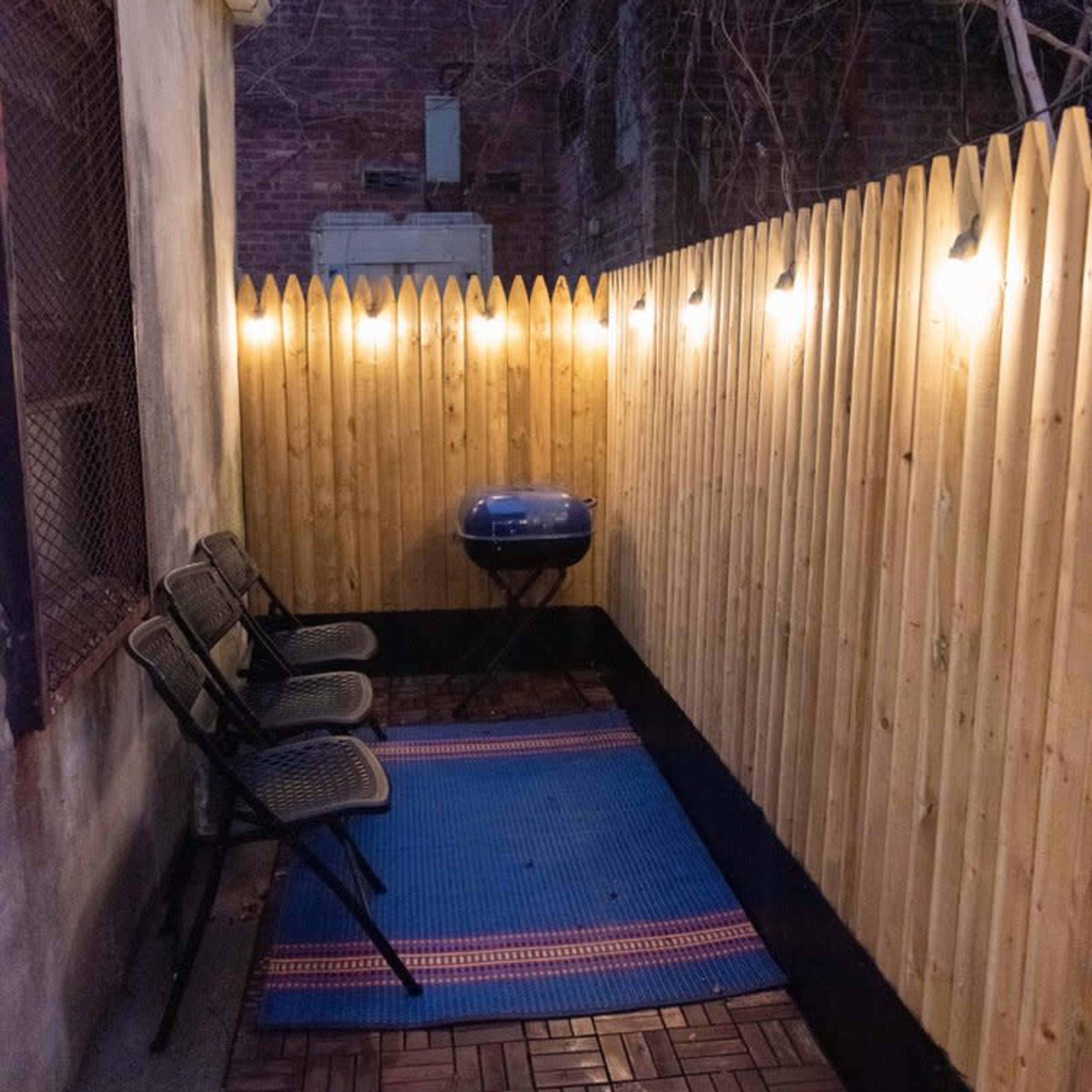 A narrow outdoor space with a wooden fence, two black chairs, a blue grill, and string lights overhead.
