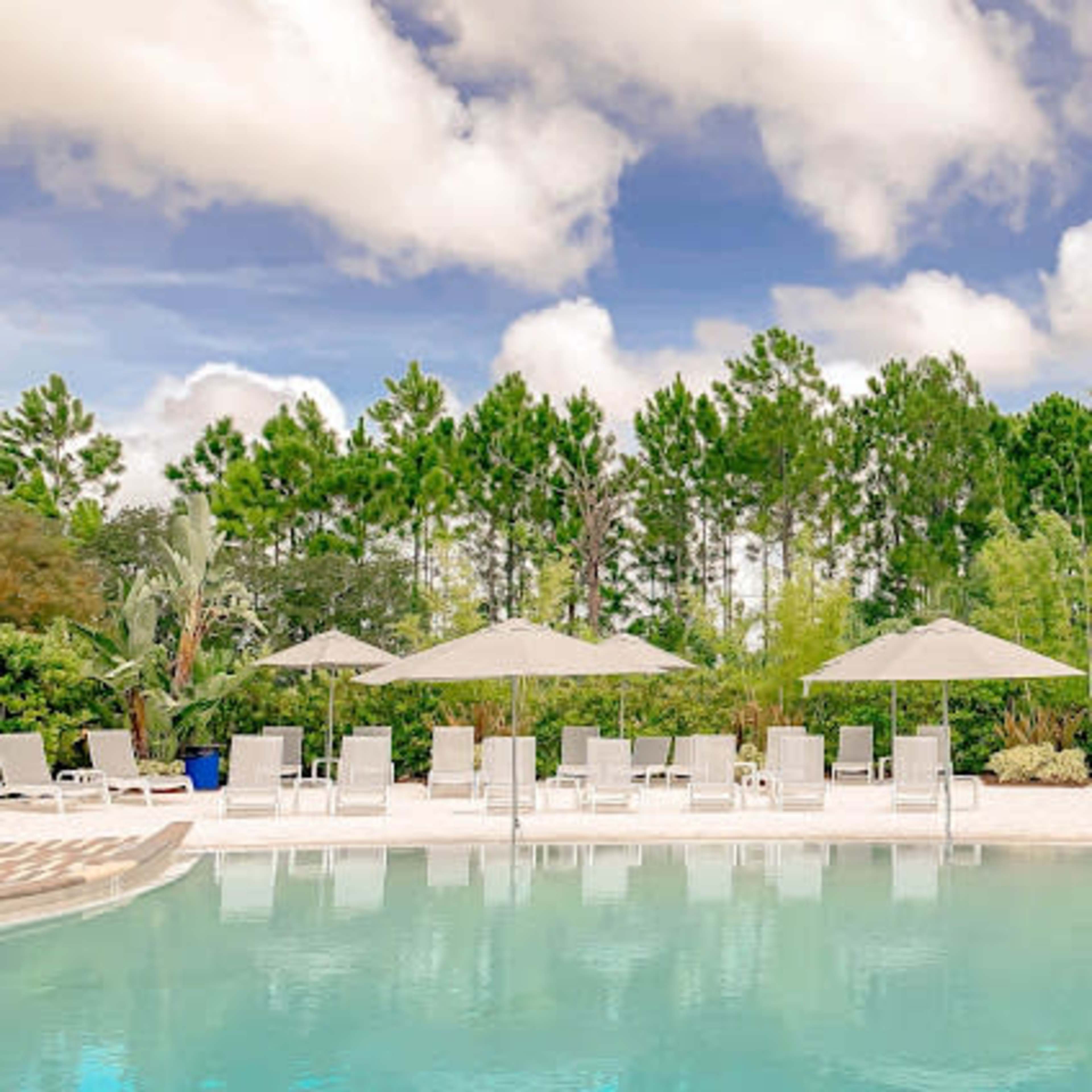A clear swimming pool is surrounded by sun loungers and shaded umbrellas, with lush greenery in the background.