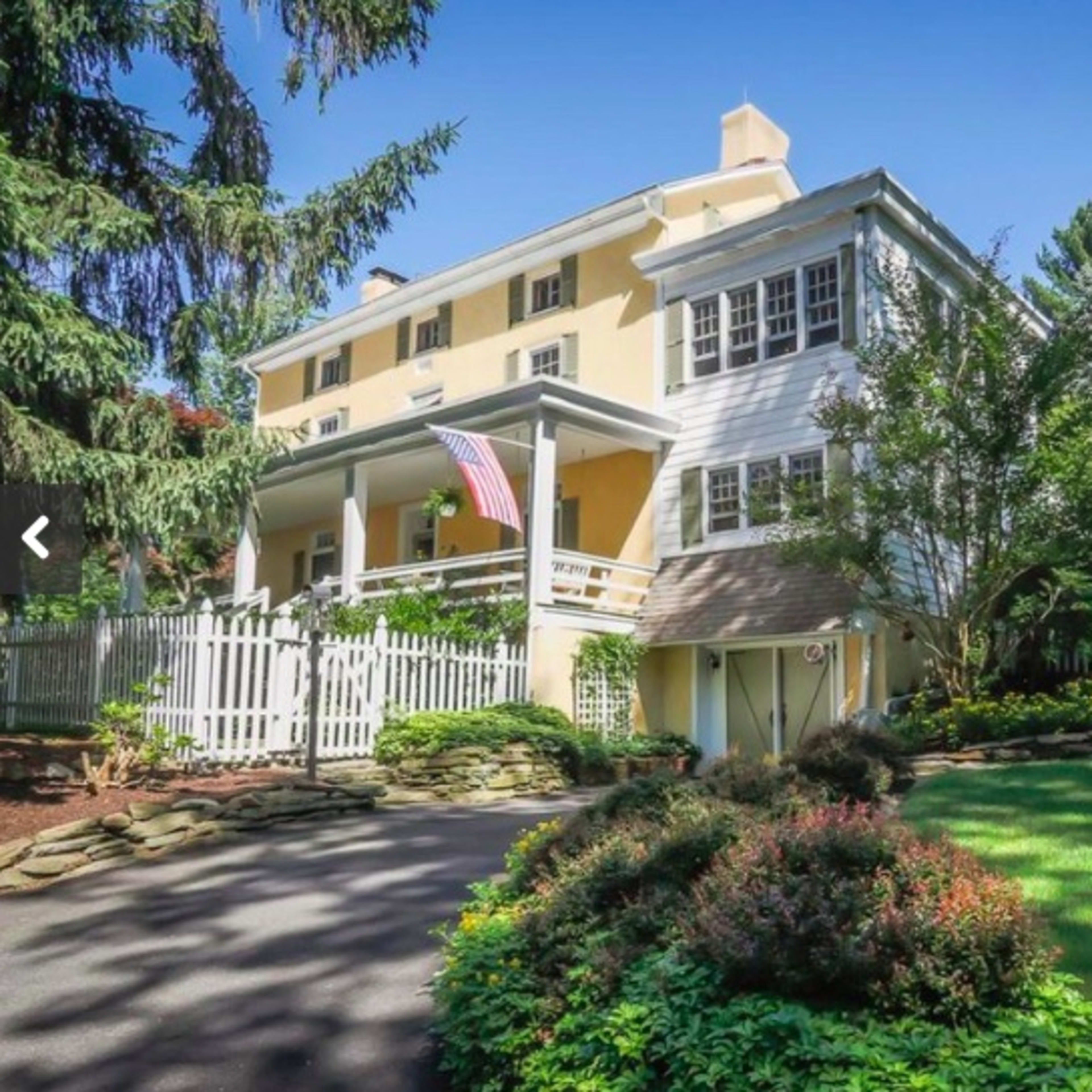 A two-story house with a yellow exterior and a front porch, surrounded by greenery and a white picket fence.