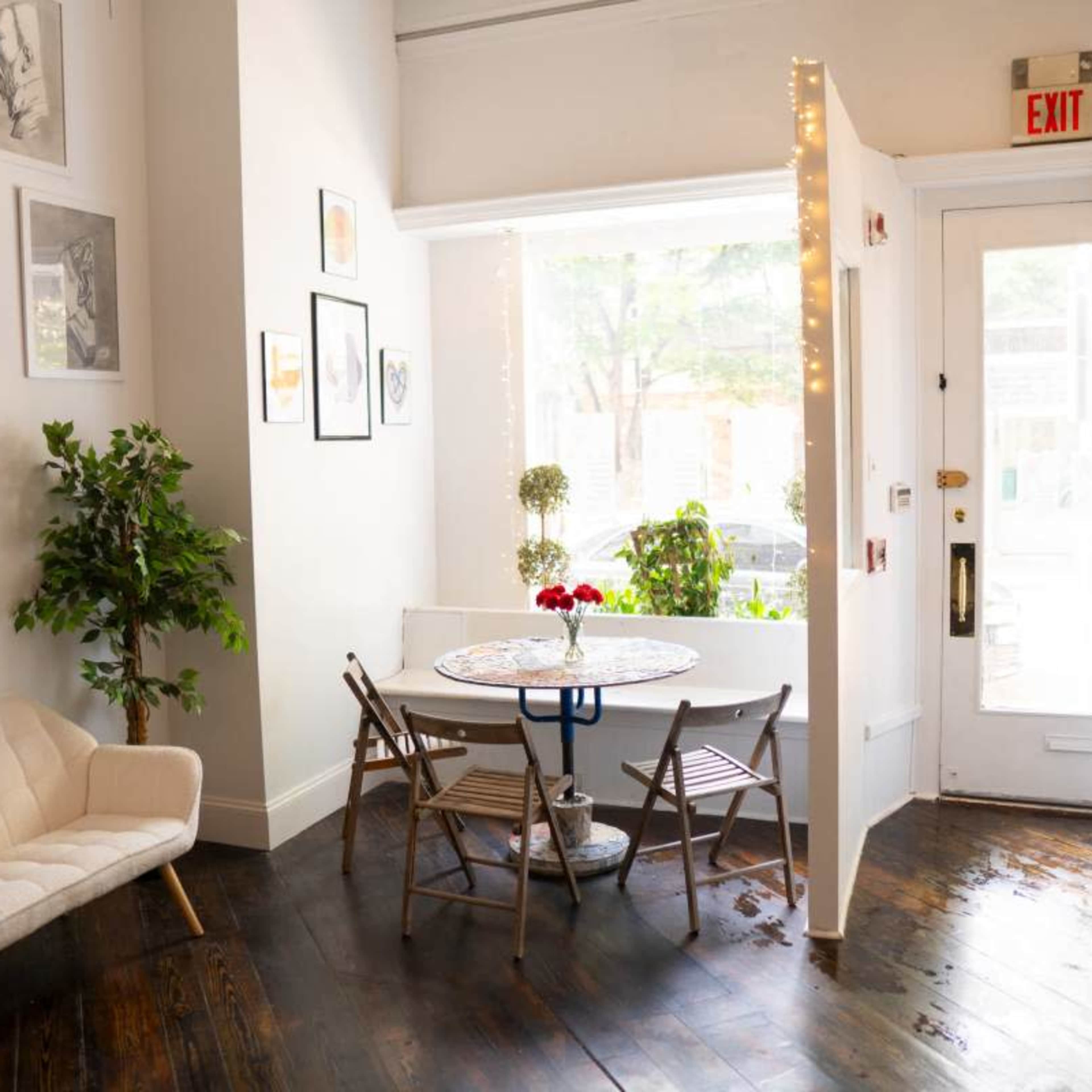 A small, bright room featuring a seating area with a light-colored chair and a round table adorned with a vase of red flowers, beside a window.