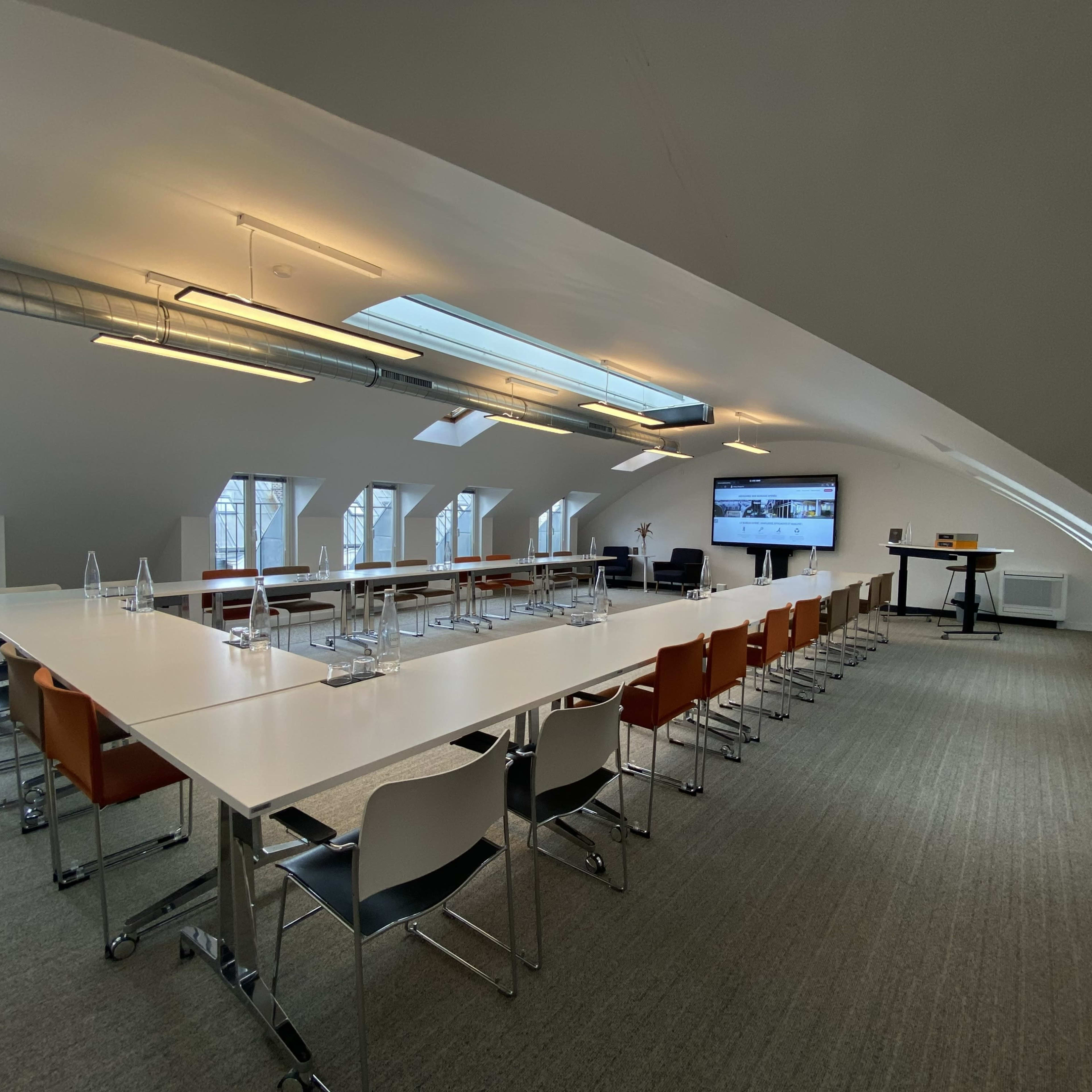 A spacious meeting room features a long table with chairs, a large screen, and skylights illuminating the area.
