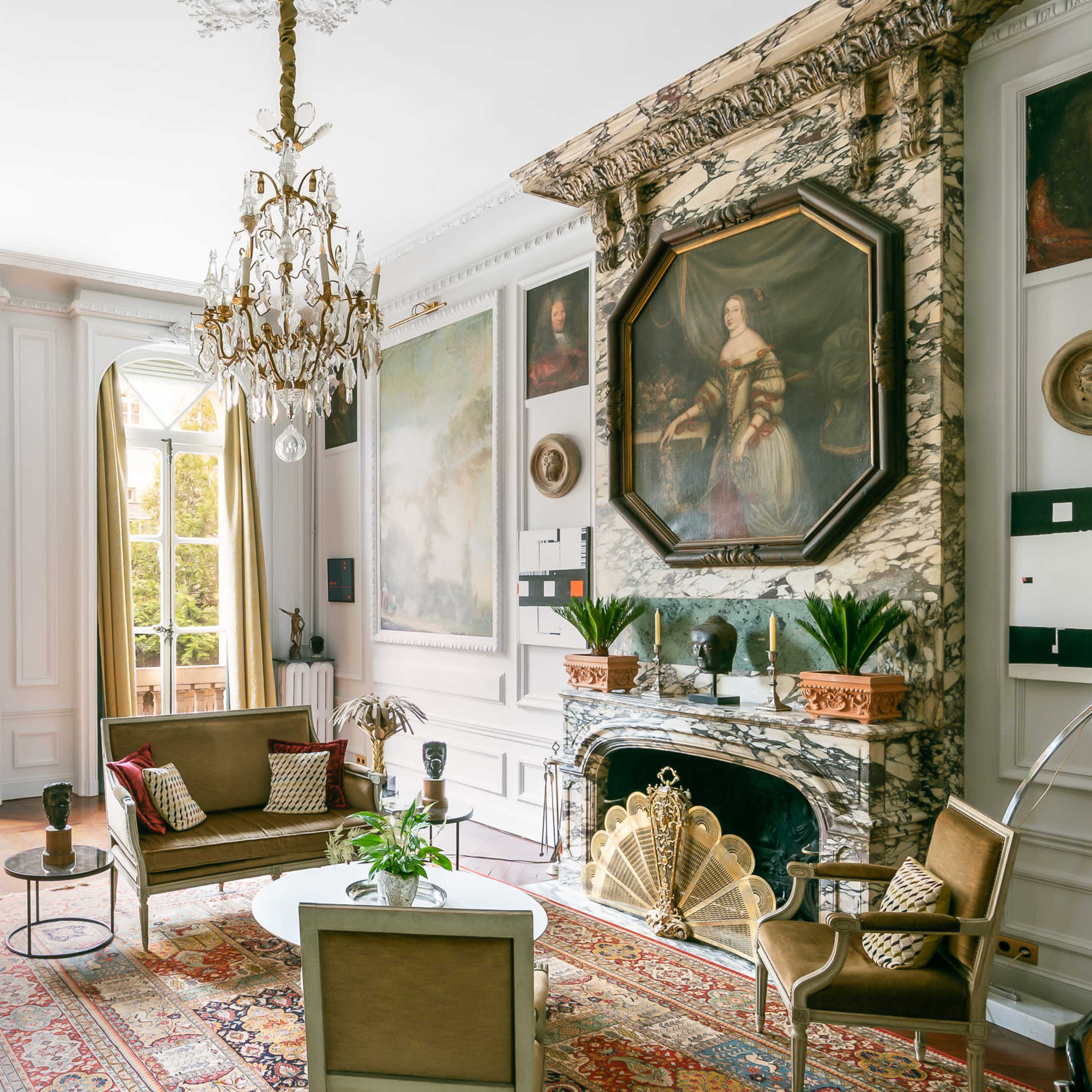 An elegantly furnished living room featuring a chandelier, marble fireplace, artwork on the walls, and large windows that open to a garden view.