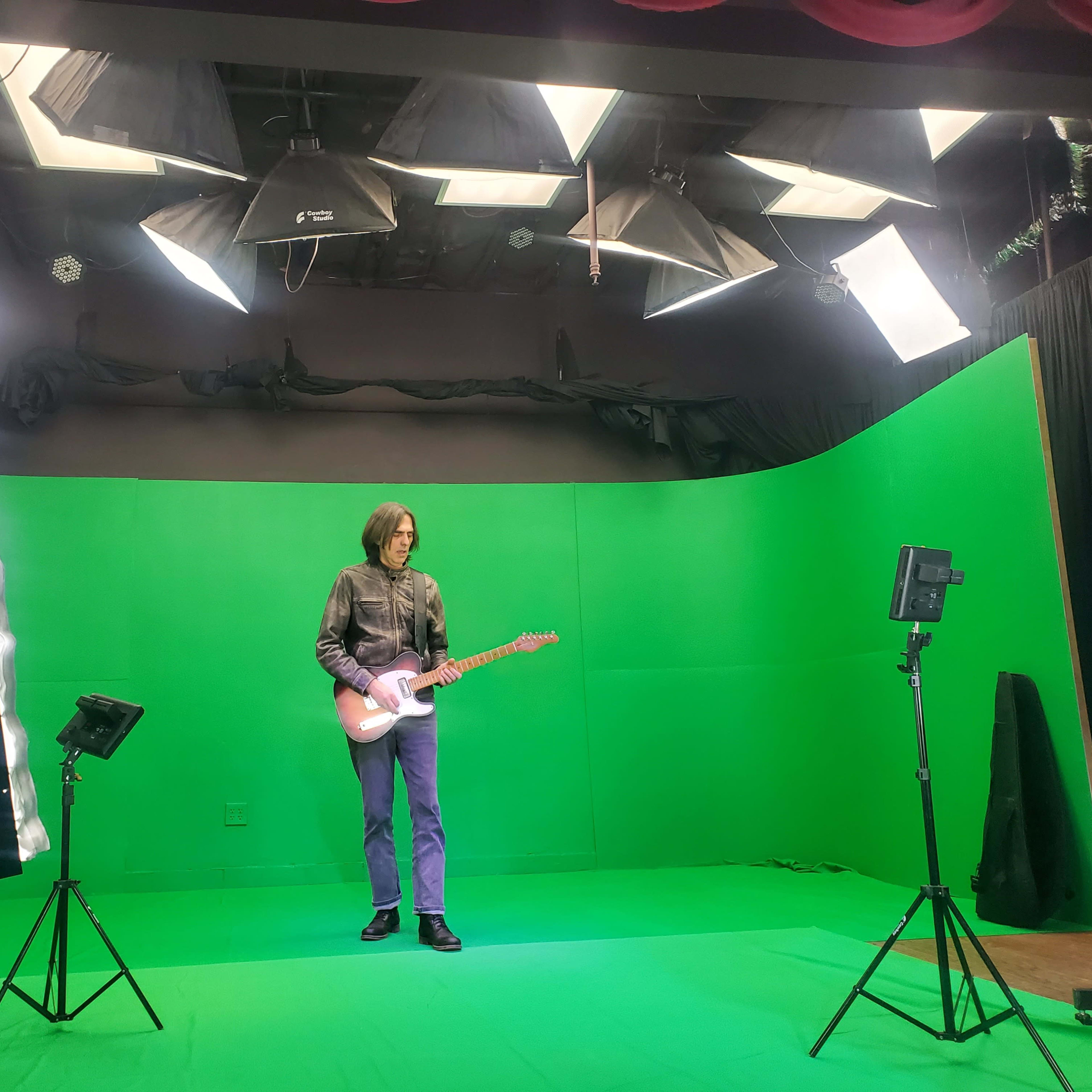 A person stands on a green backdrop set, holding an electric guitar, surrounded by studio lighting equipment.