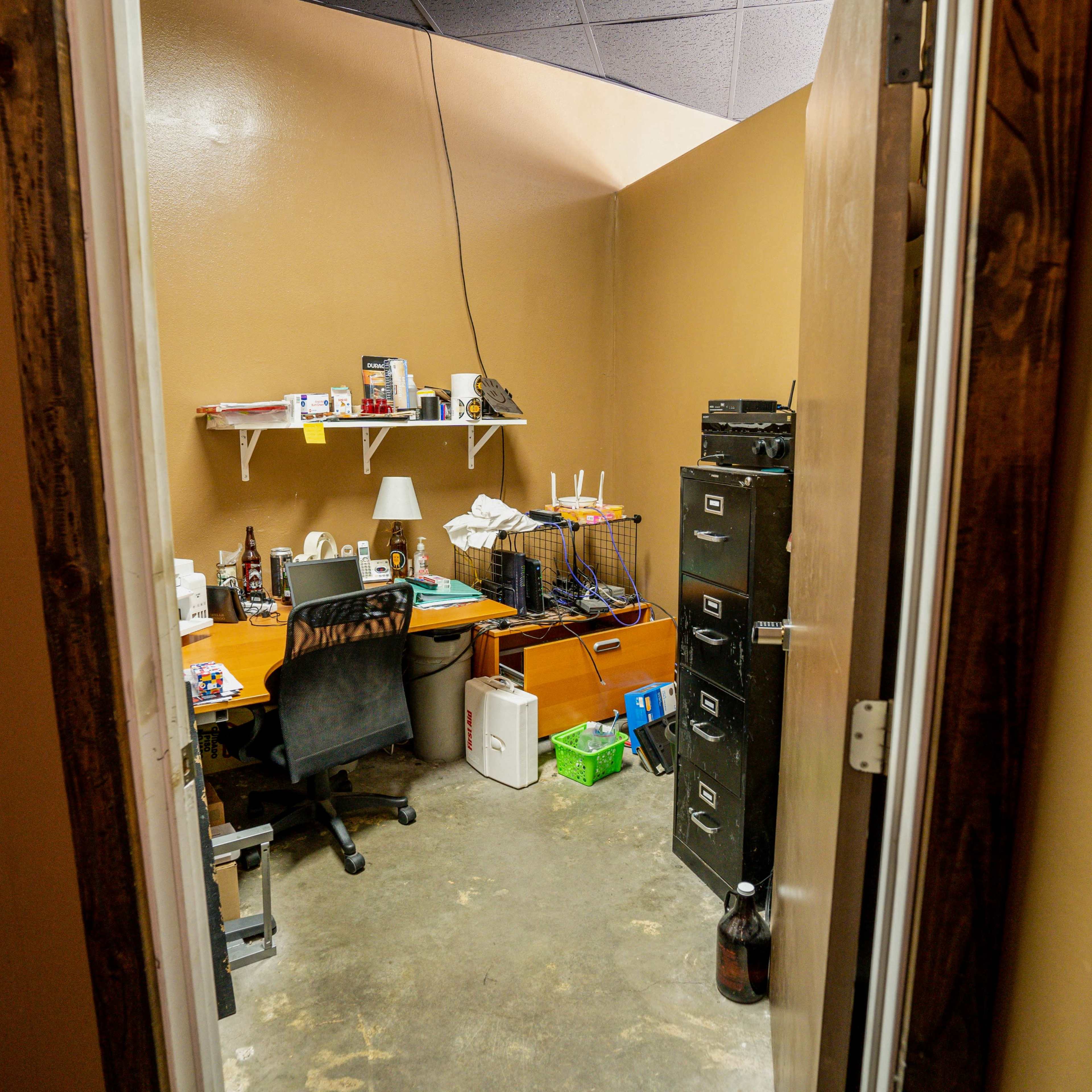 A small office area viewed through an open door, featuring a desk, filing cabinets, and shelves with various items.