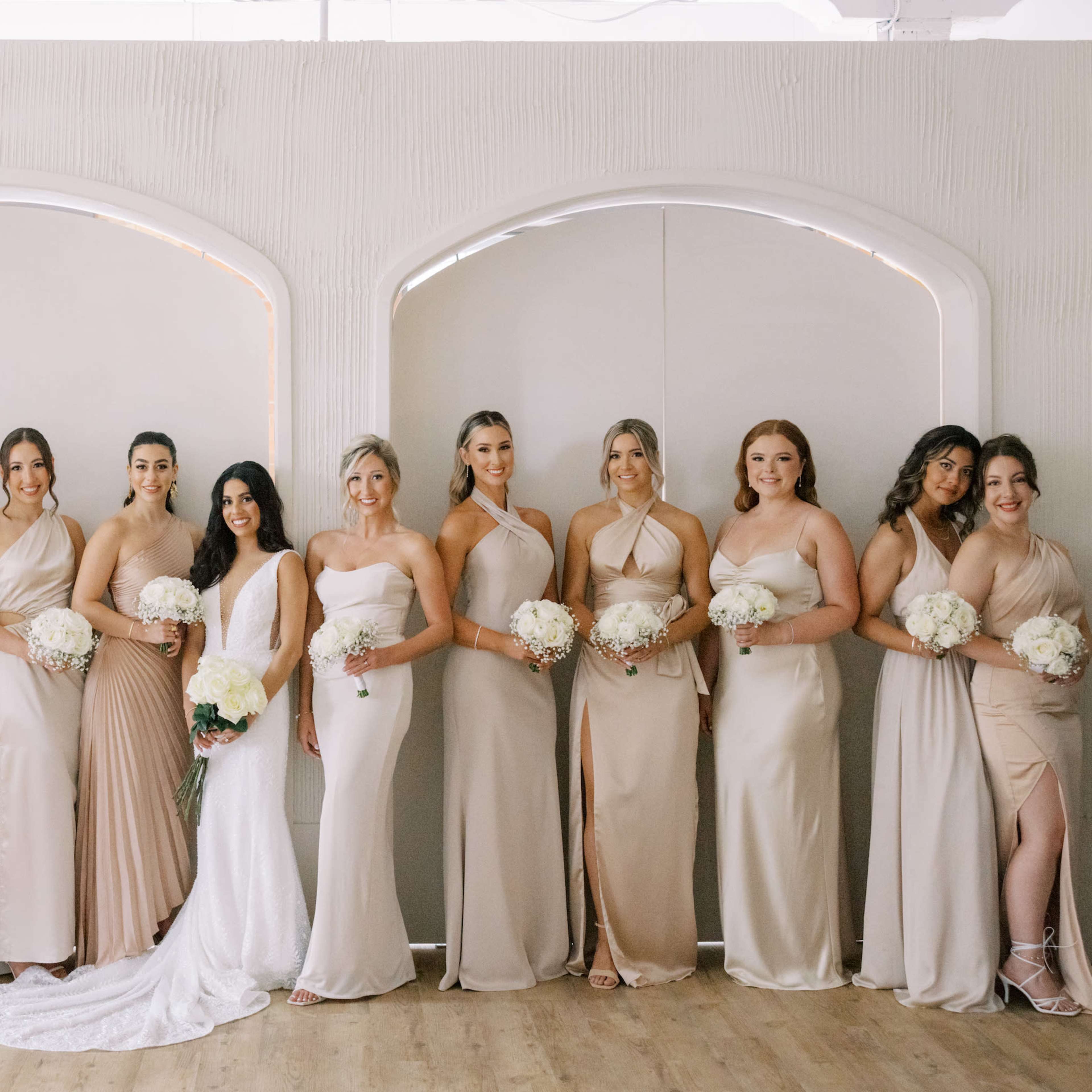 A group of fifteen women in coordinated dresses poses together with floral bouquets in a well-lit indoor setting.