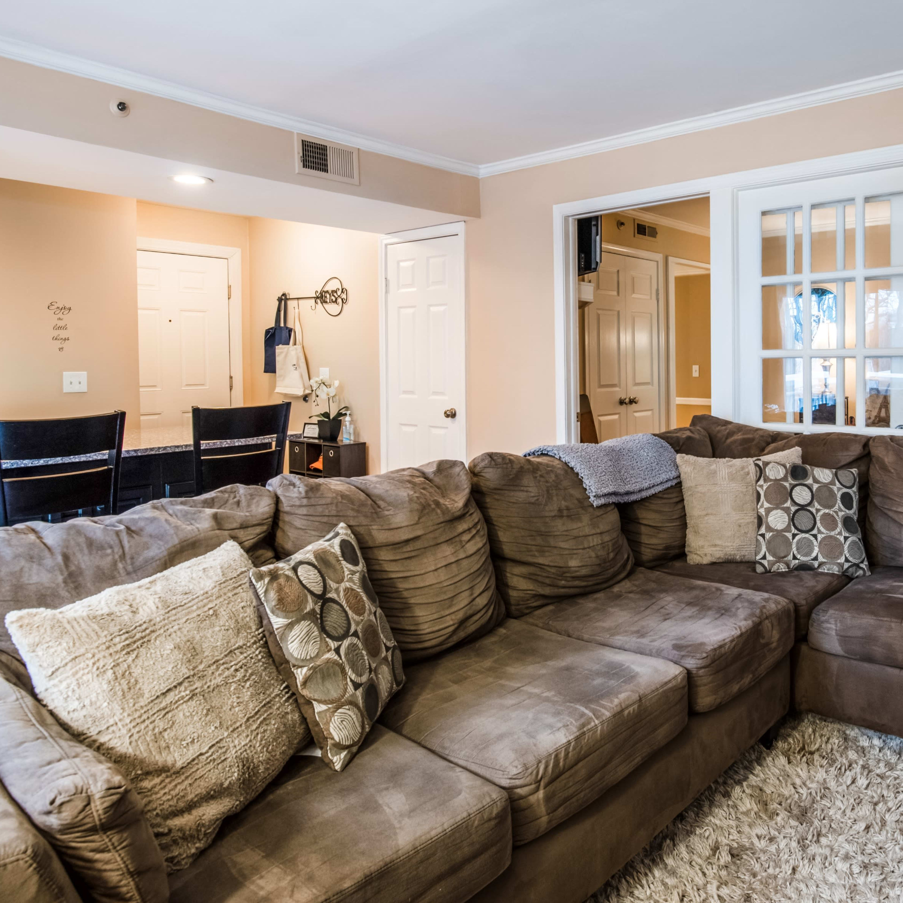 A spacious living area featuring a large, sectional brown couch with decorative pillows, next to a dining area and kitchen visible through a set of French doors.