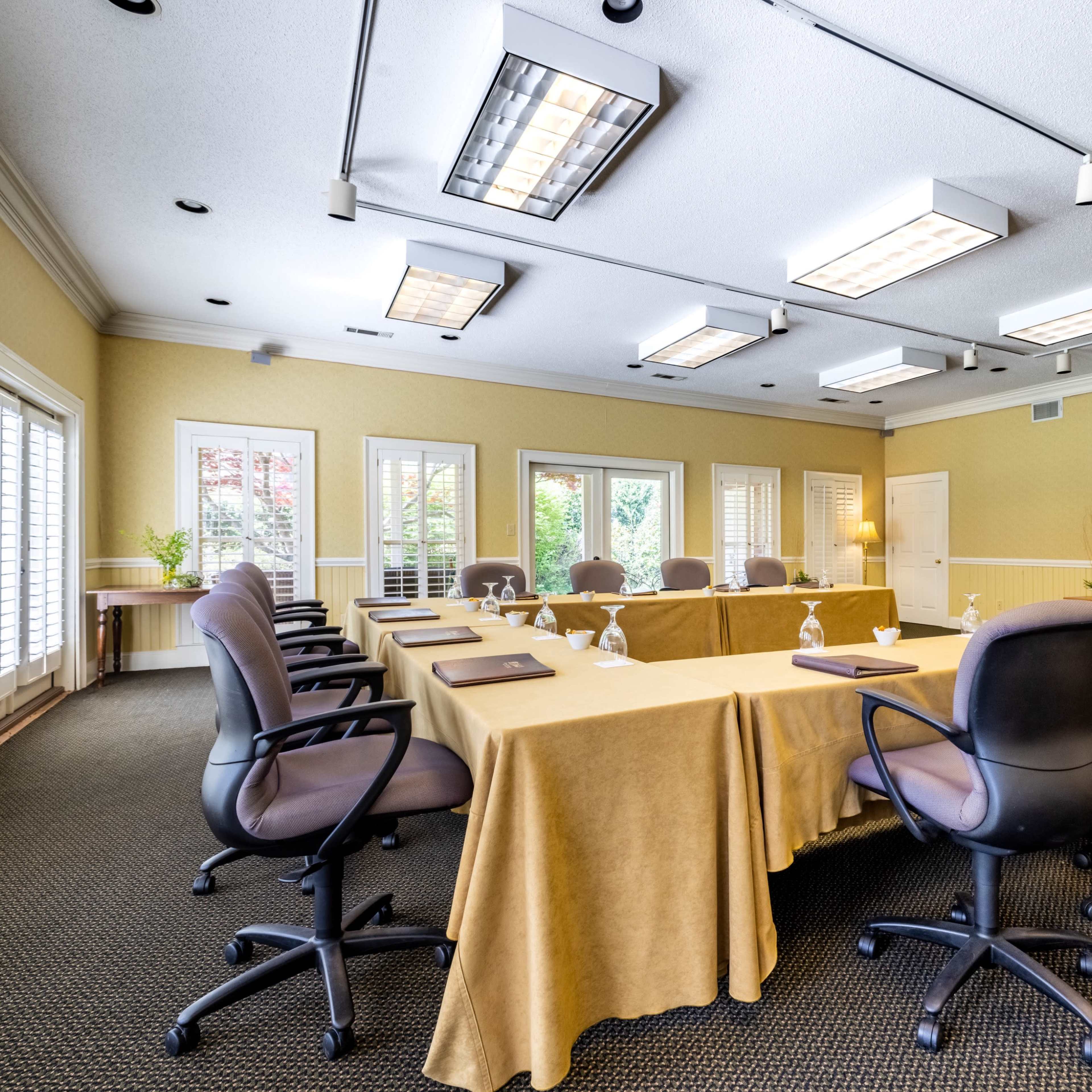 A conference room features a rectangular table surrounded by rolling chairs, with large windows allowing natural light and a screen at one end of the room.