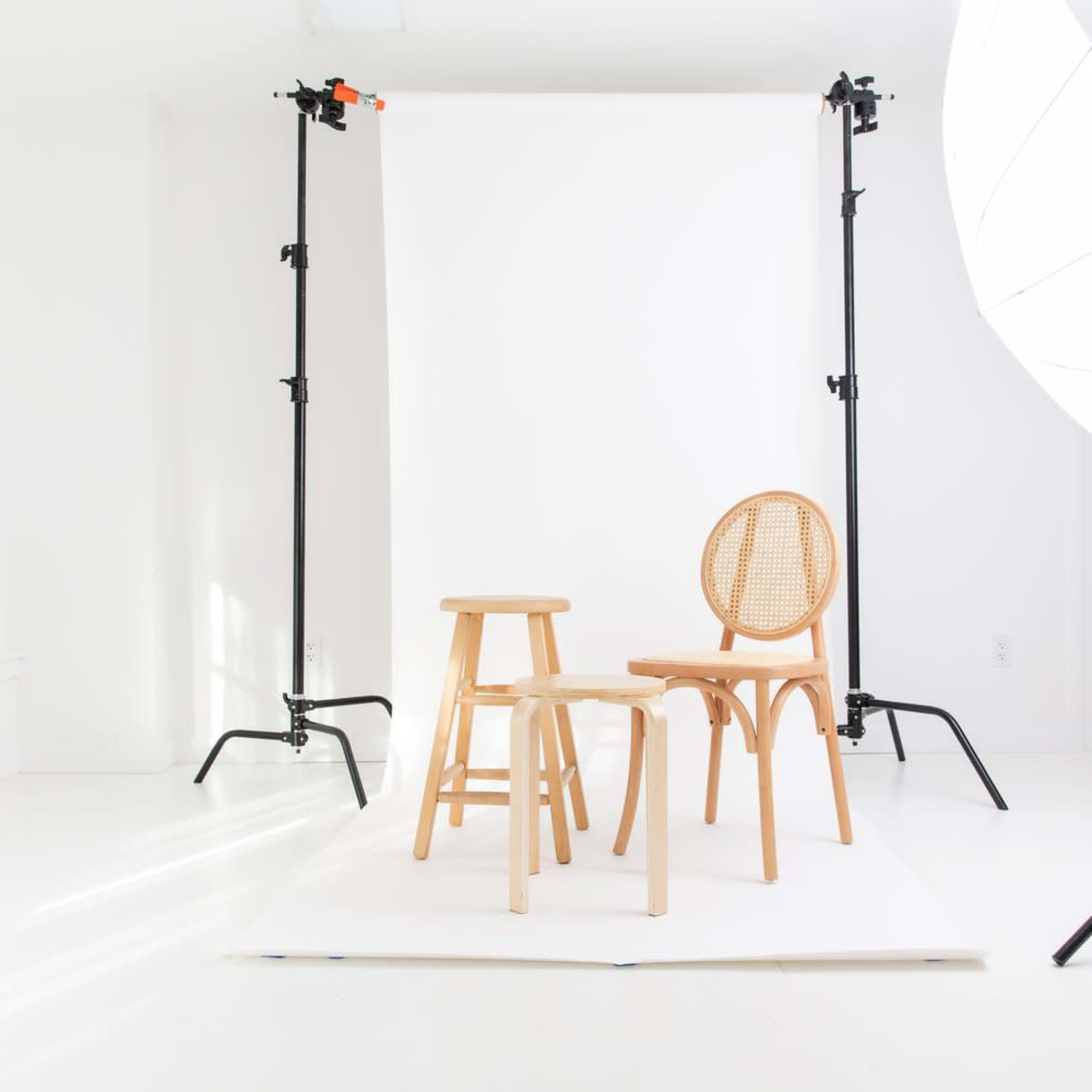 A bright, minimalist photography studio setup with two wooden chairs and a stool positioned in front of a white backdrop.