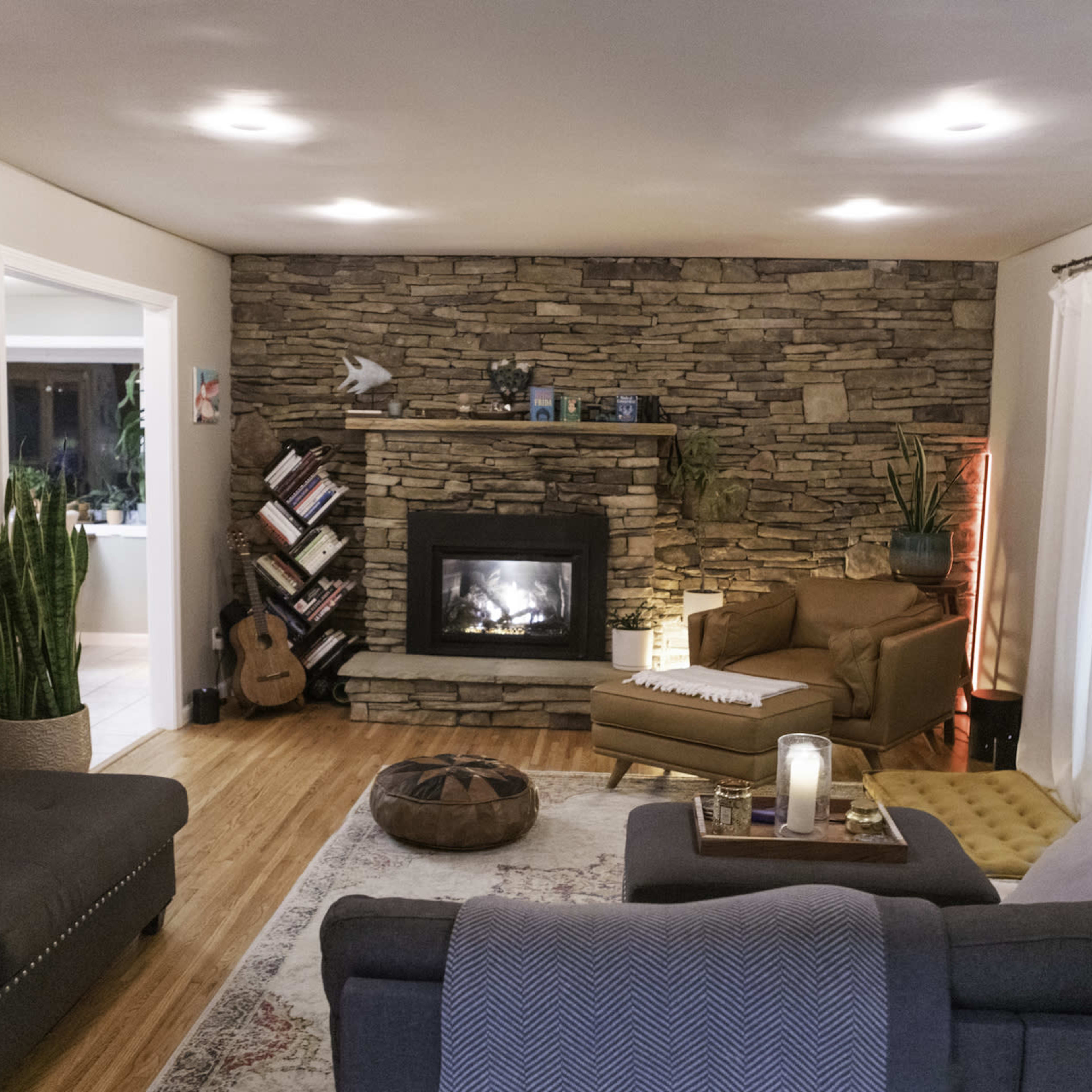 The living room features a stone fireplace surrounded by plants, comfortable seating, and a mix of decorative elements including artwork and a cozy area rug.