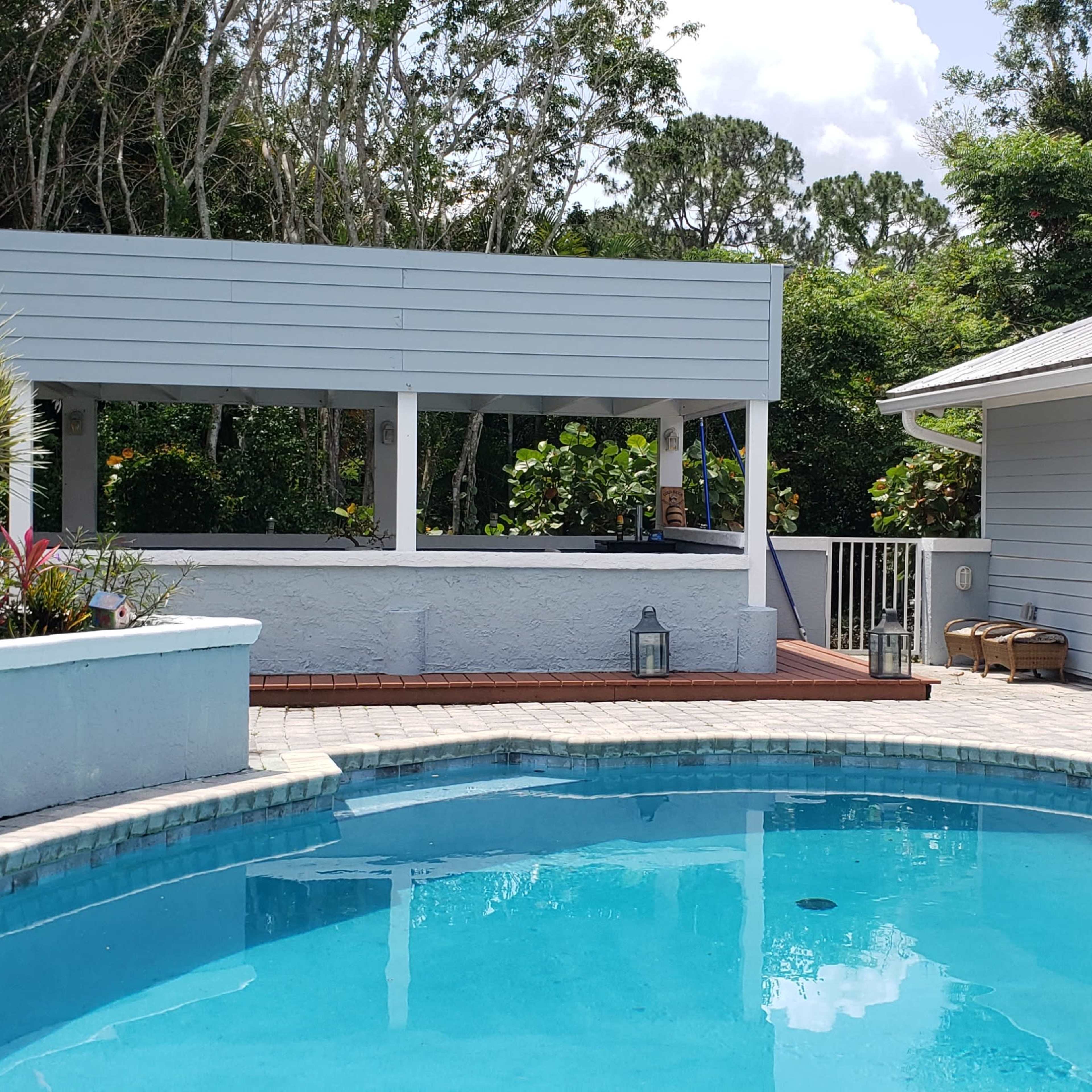 A backyard with a swimming pool surrounded by a paved deck, a wooden terrace housing plants, and a light-colored house on the right.