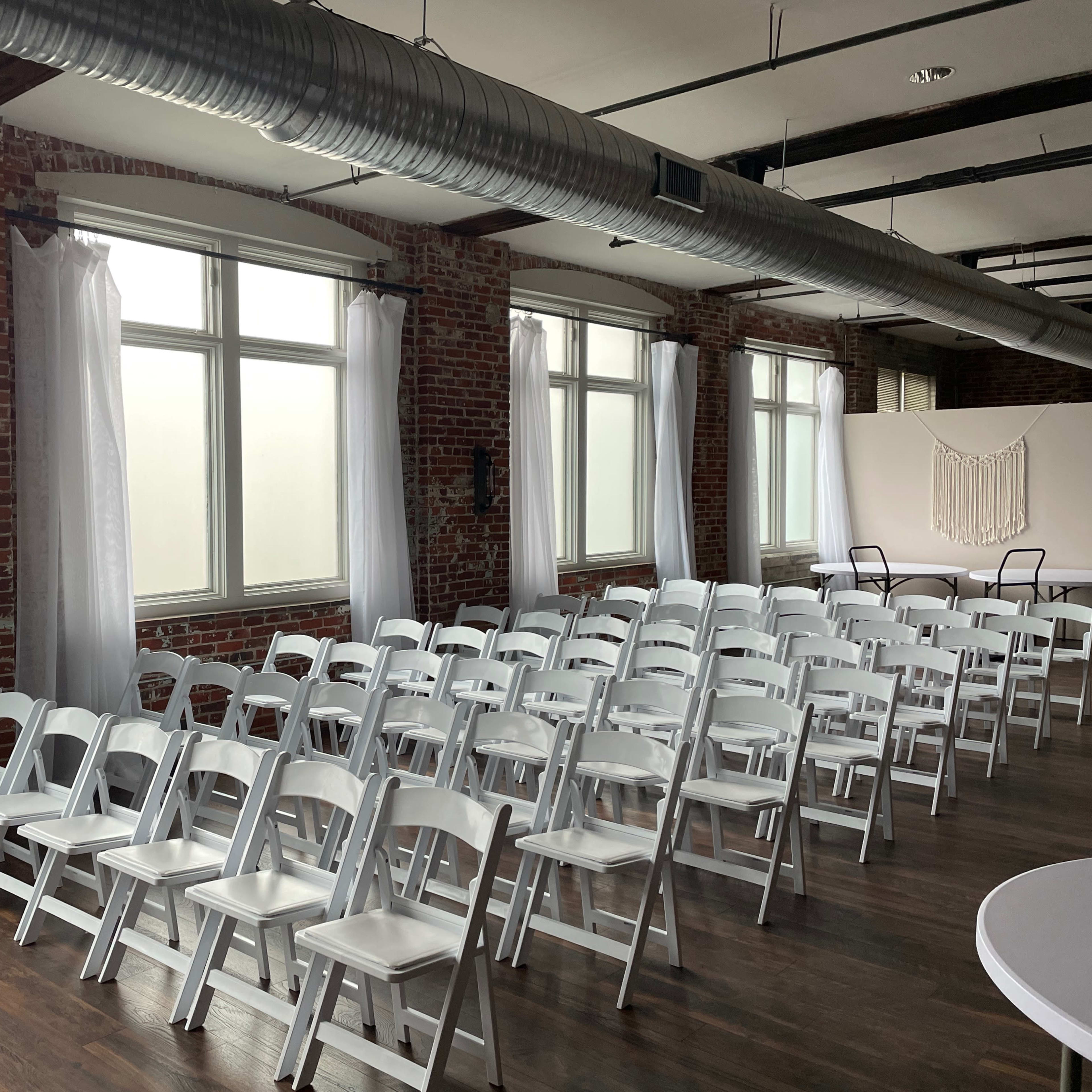 A row of white folding chairs is arranged in front of large windows in a room with exposed brick walls.