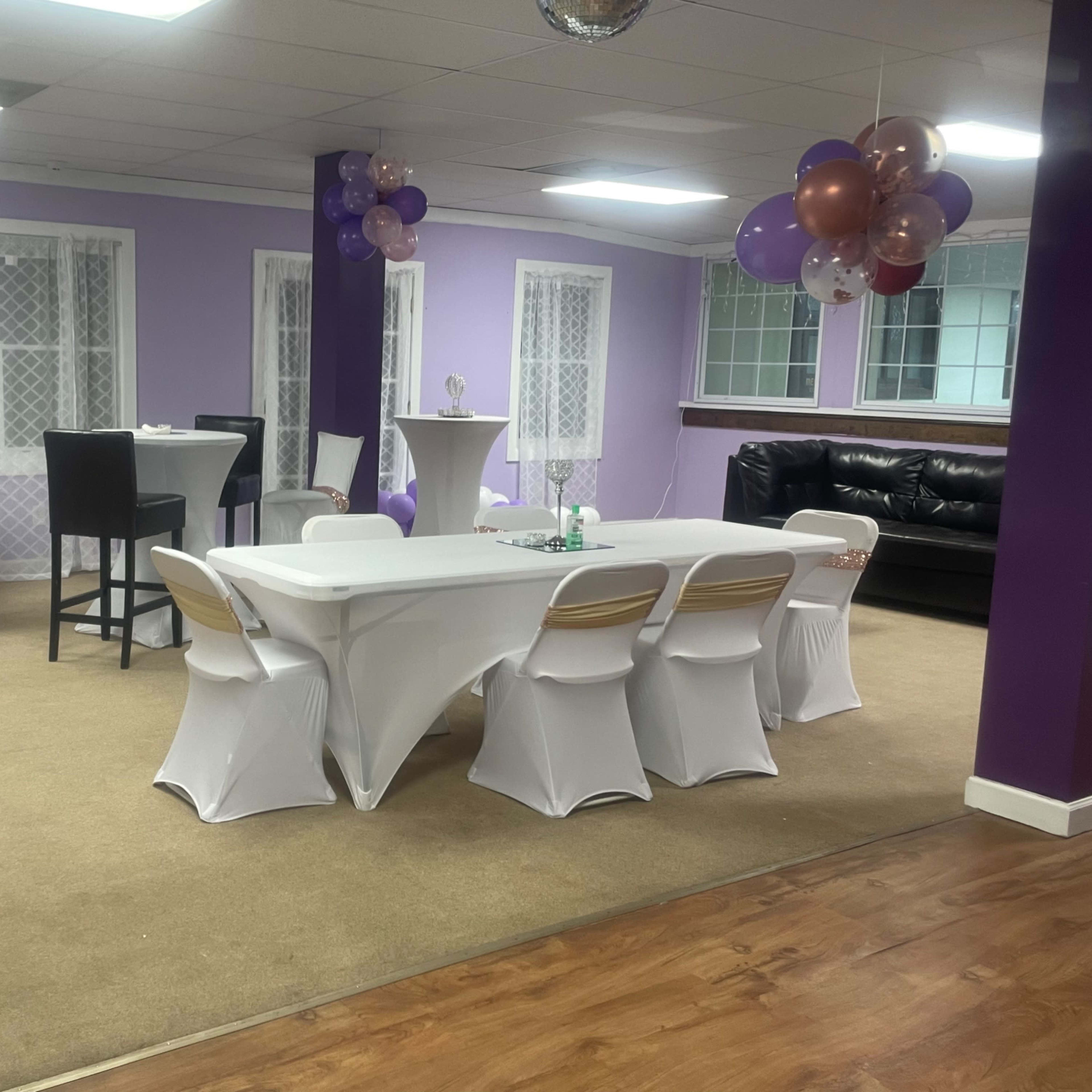 A party venue with purple walls, tables covered in white tablecloths, and decorative balloons in the corner.