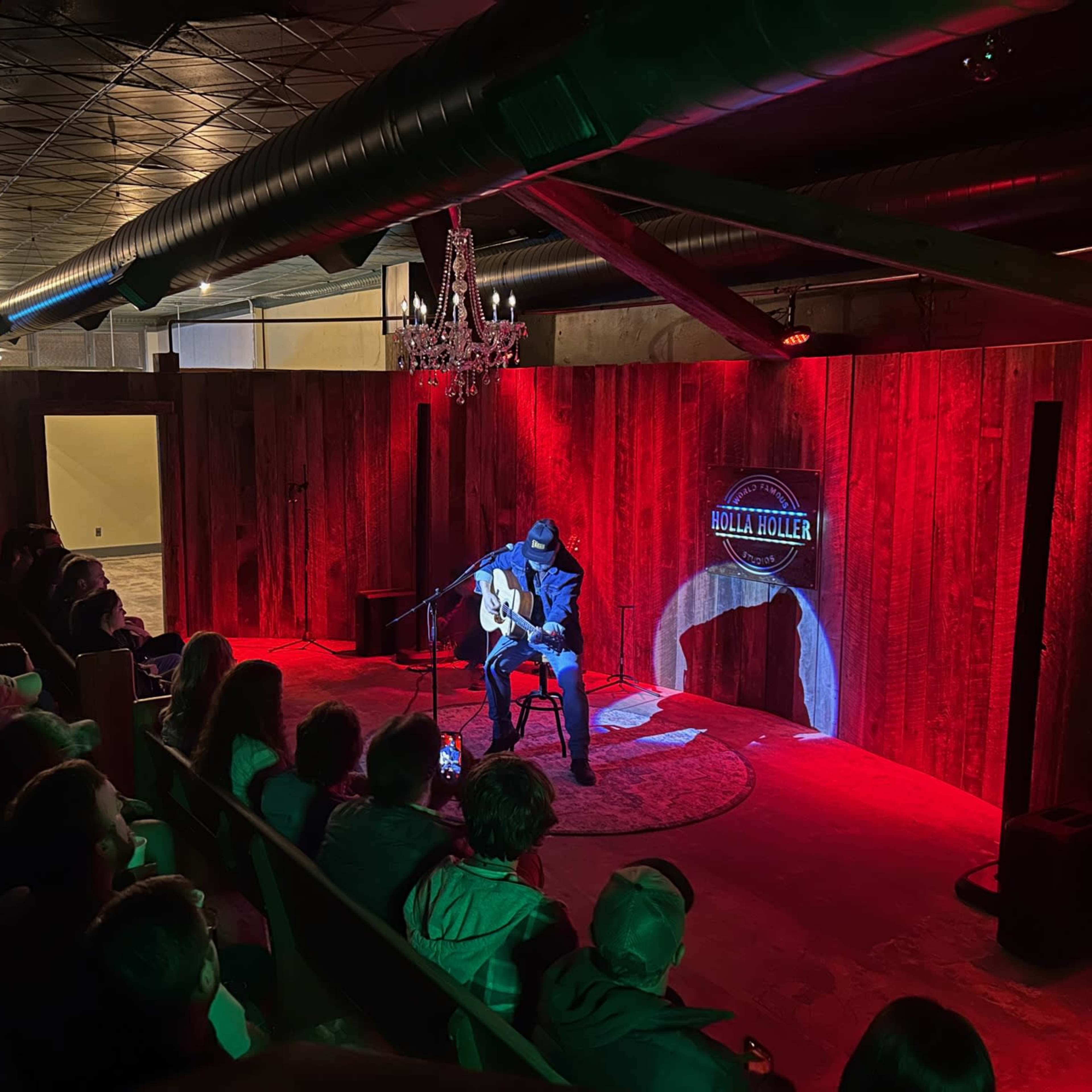 A performer plays guitar on stage in a dimly lit venue while an audience watches from the seats.