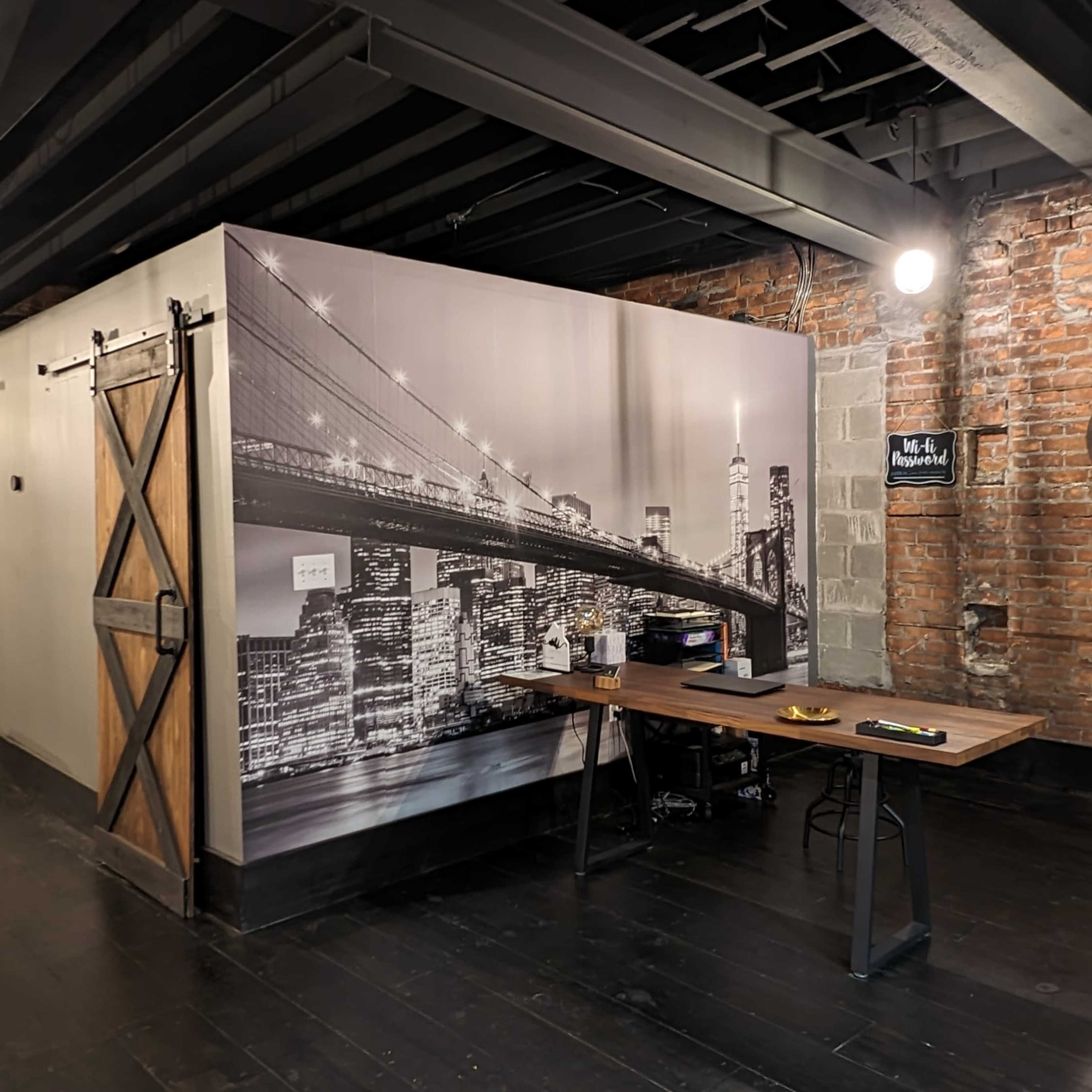 An interior space featuring a decorative wall mural of a bridge, a wooden table with items on it, exposed brick walls, and industrial lighting fixtures.