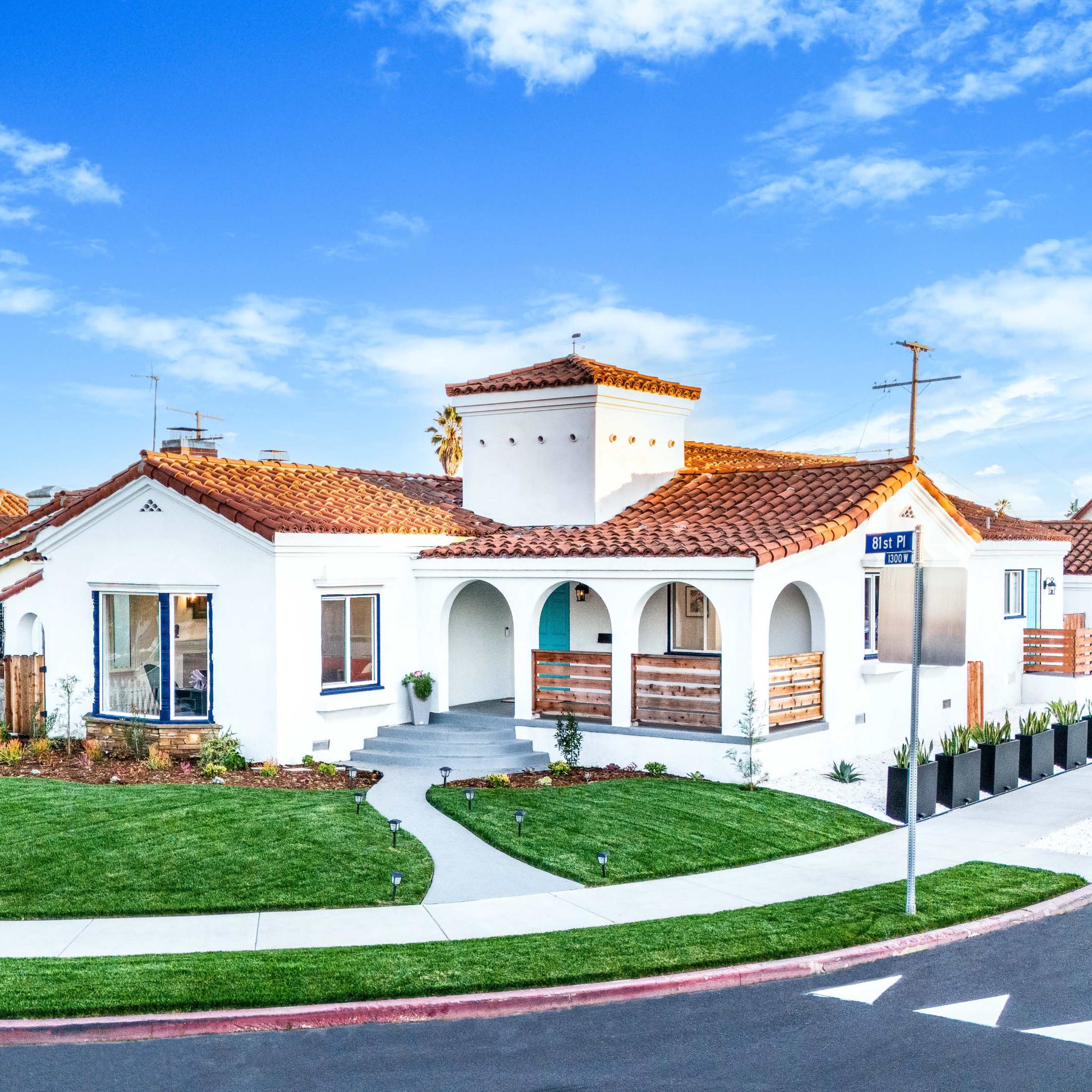 A white stucco house with a red tile roof is situated on a corner lot, surrounded by green lawns and flower beds.