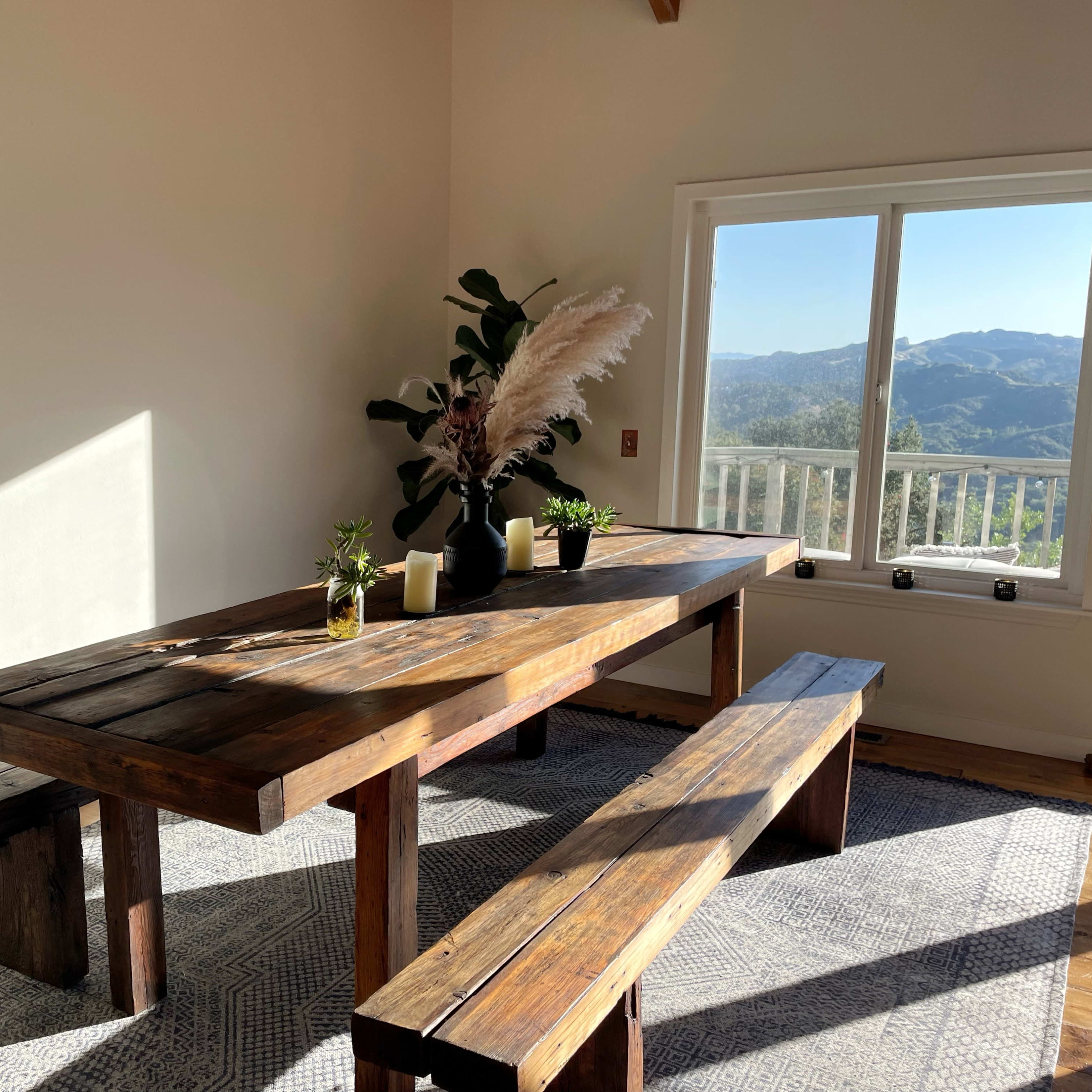 A wooden dining table with benches is set in a sunlit room, beside a large window offering a view of the mountains.