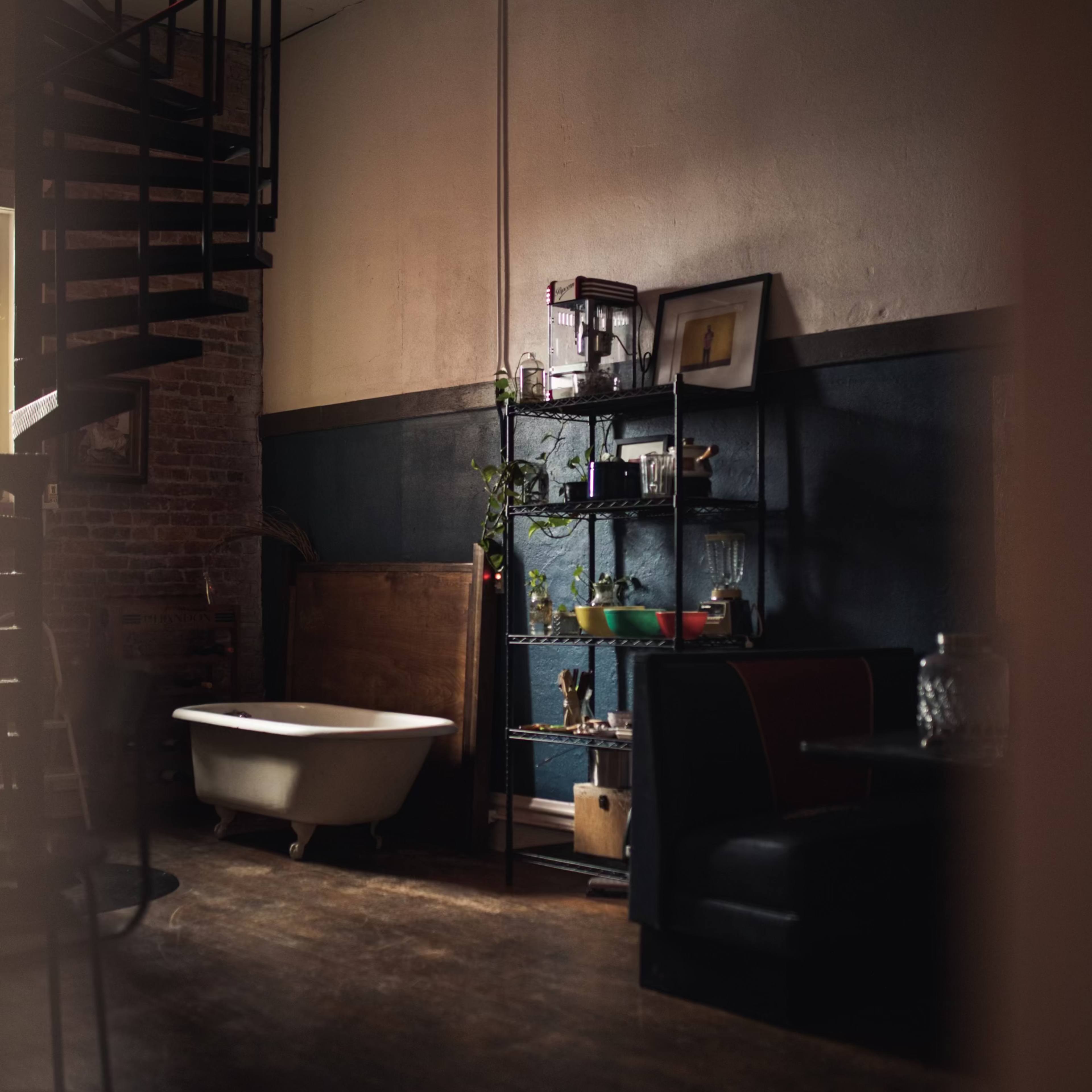 A dimly lit interior with a spiral staircase, a vintage bathtub, and a shelf displaying various decorative items against a dark blue wall.
