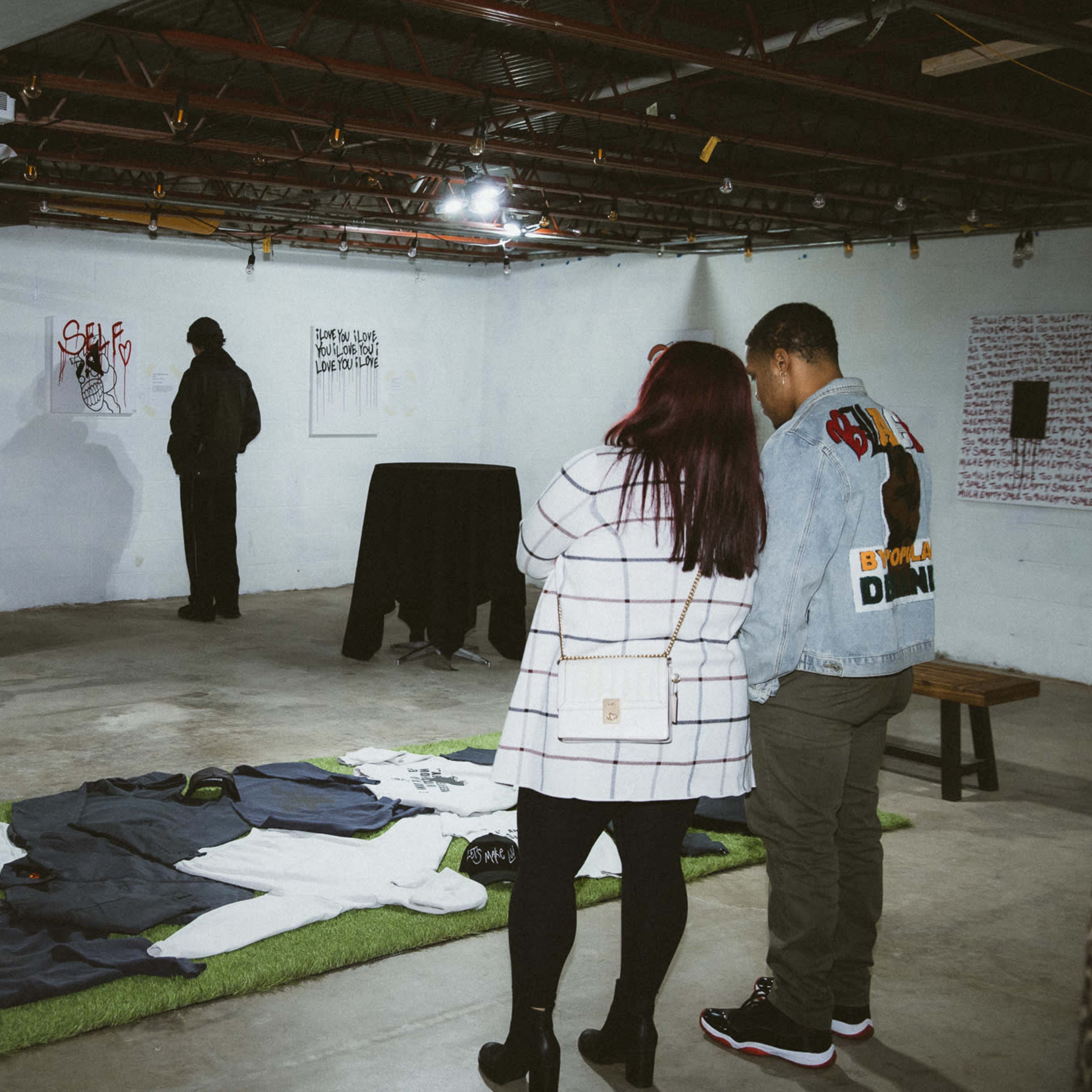 An art exhibition in a basement with two individuals examining artwork on the walls and clothing displayed on the floor.