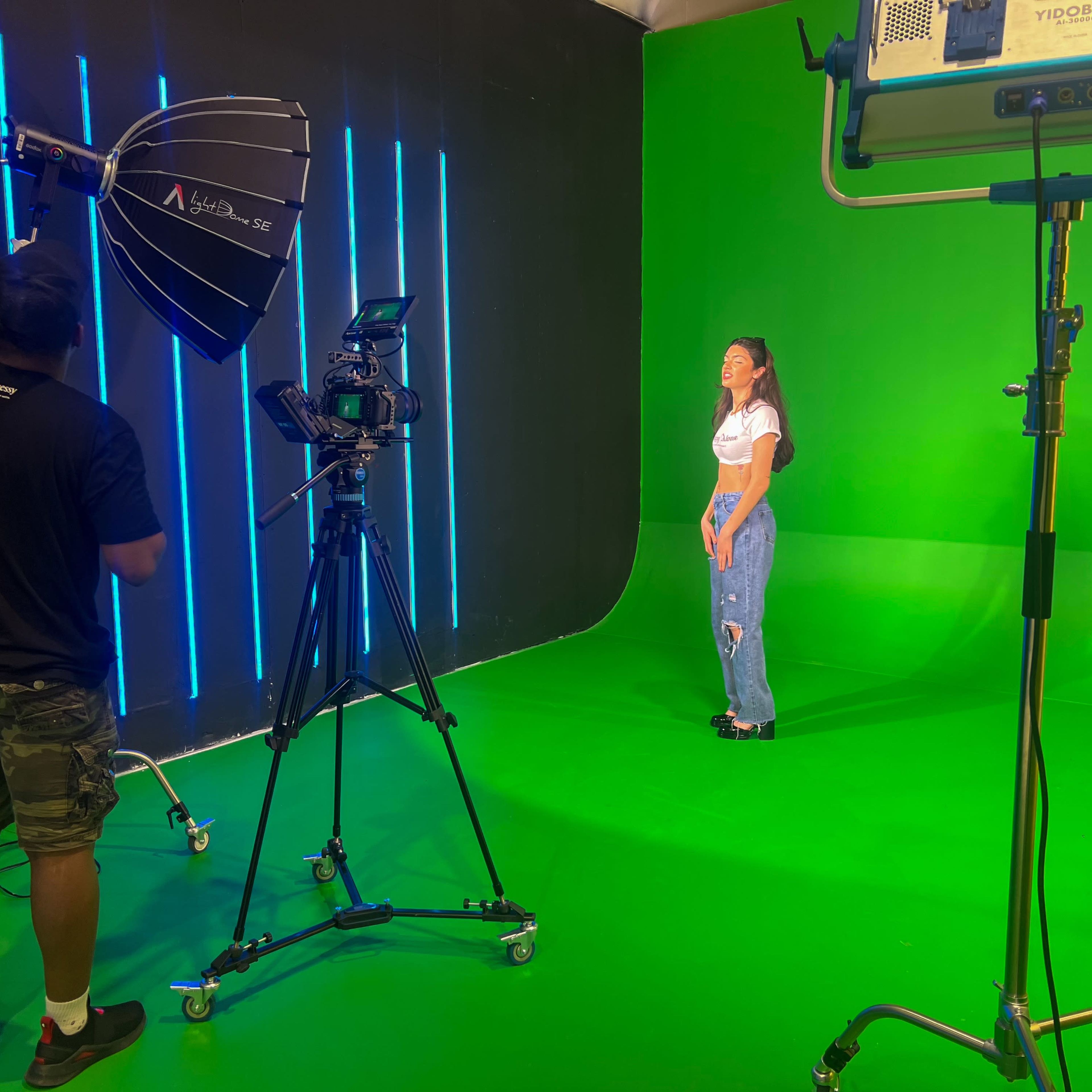 A person stands on a green screen set while another adjusts a camera and lighting equipment.