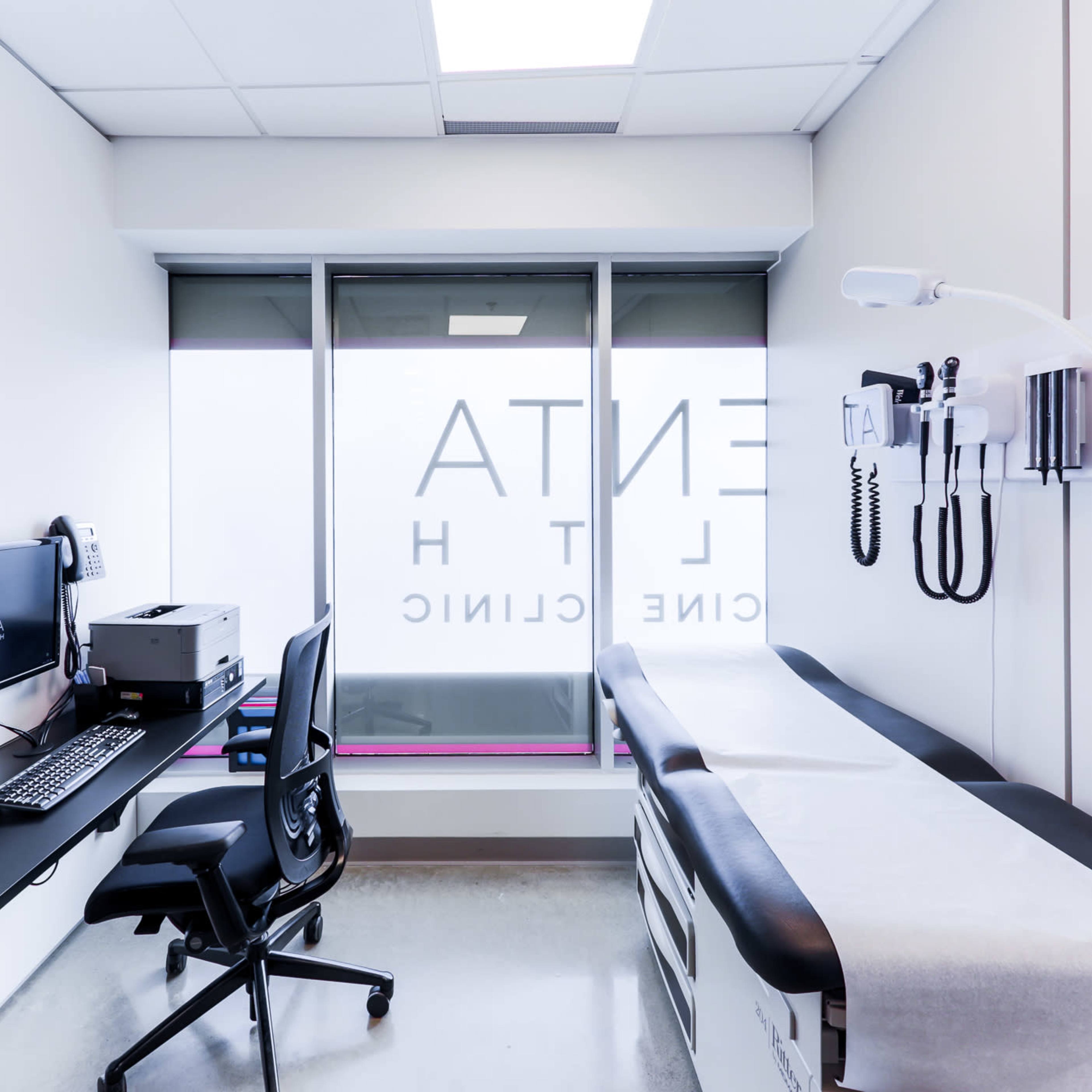 A modern medical examination room with a computer setup, a medical examination table, and medical equipment on the wall.