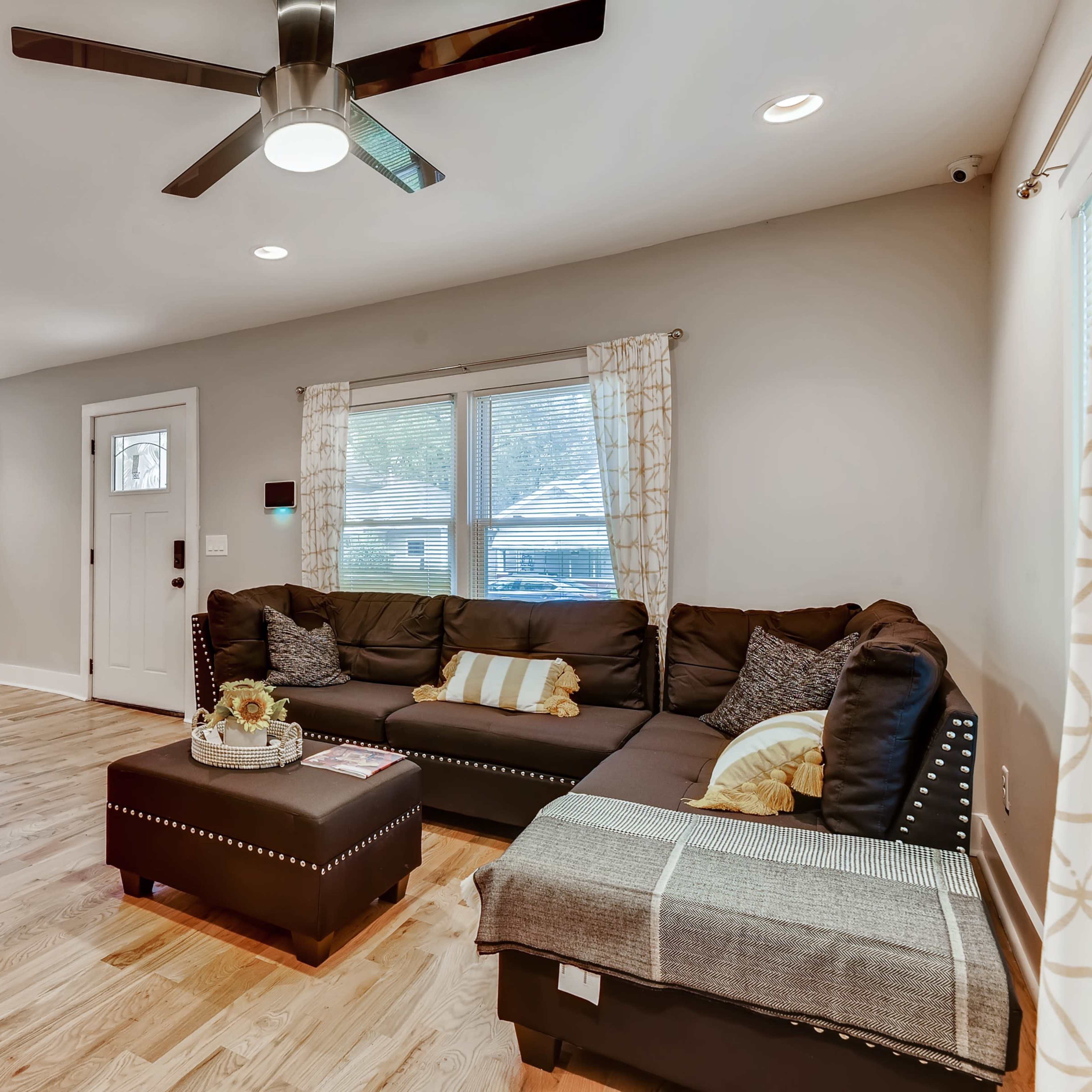 A modern living room features a sectional sofa, coffee table, and large windows allowing natural light to illuminate the space.