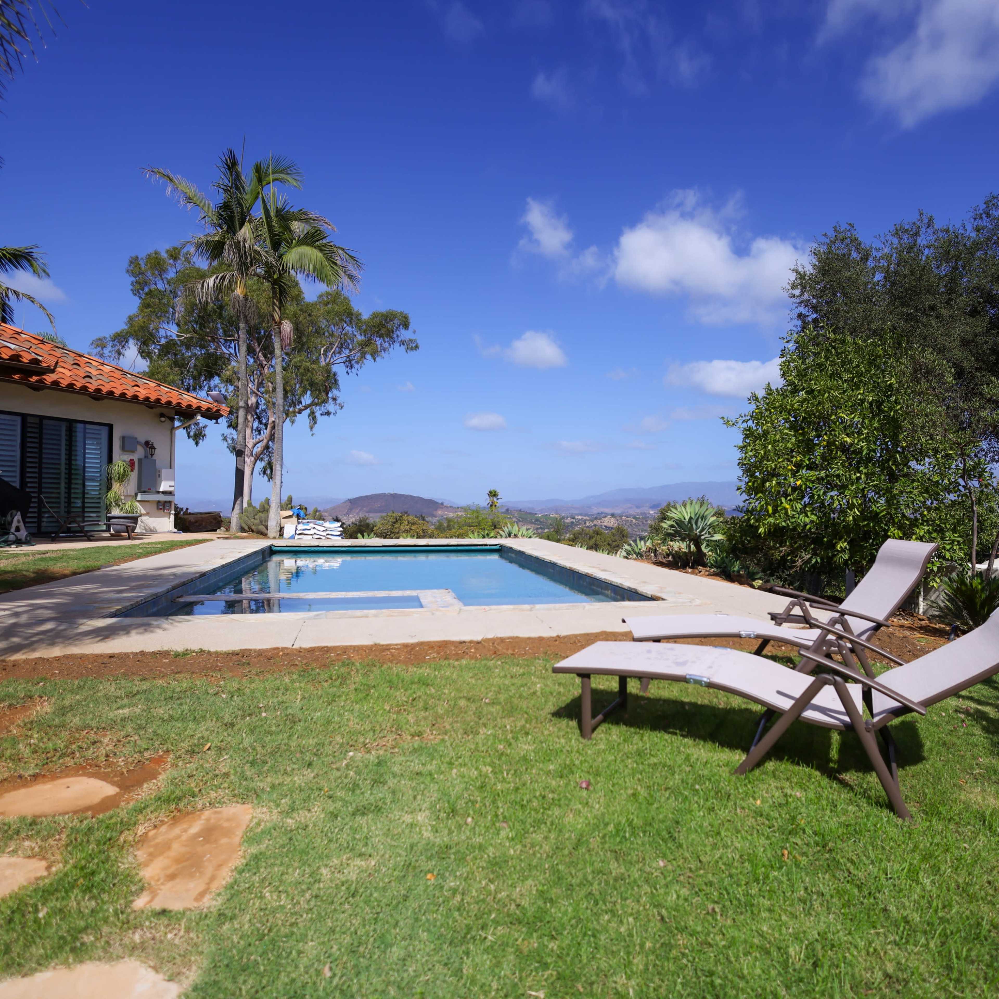 A swimming pool is surrounded by landscaped grass and patio furniture in a backyard with a view of distant hills and blue skies.
