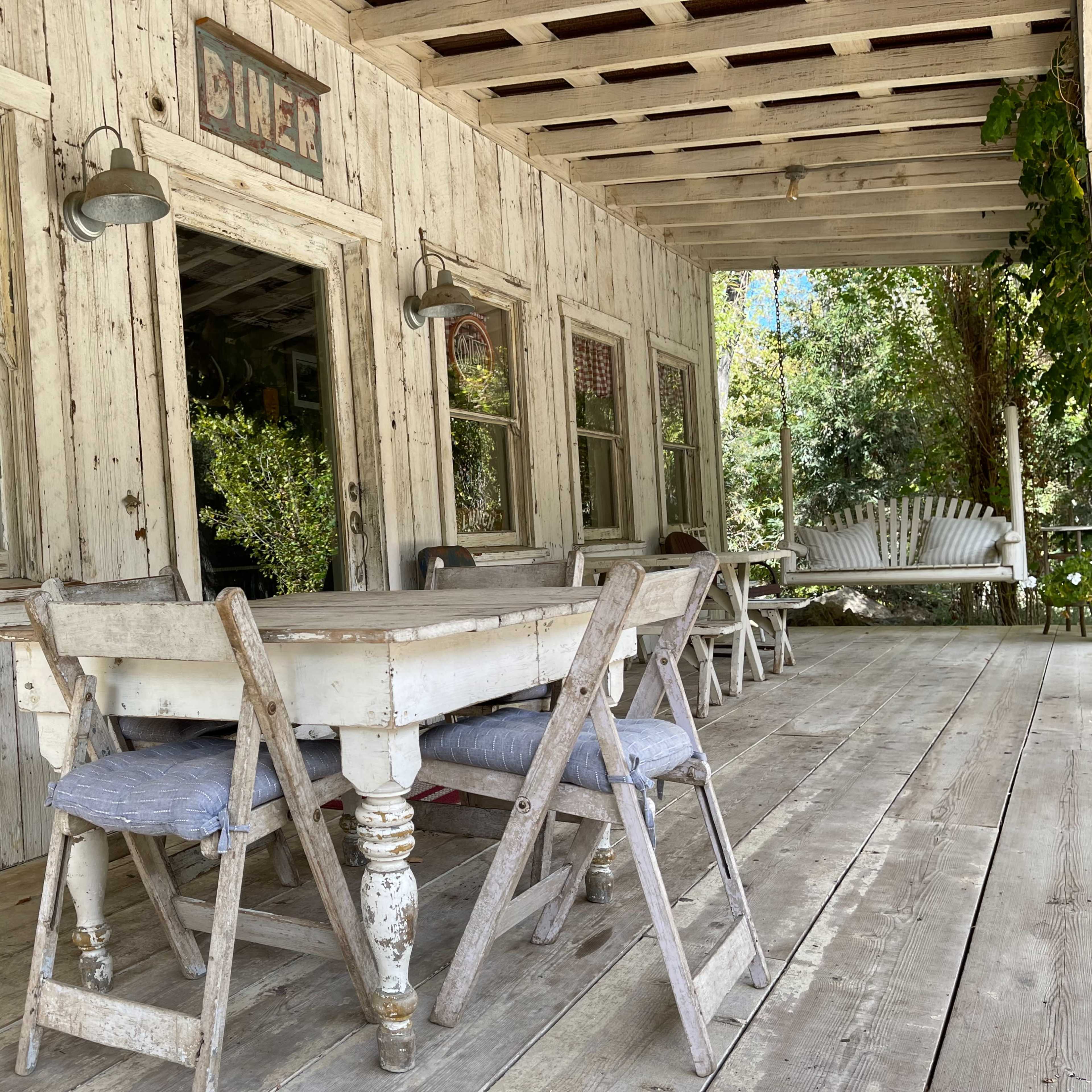 A rustic porch features a weathered wooden table and folding chairs in front of a quaint diner entrance with gingham curtains and vintage signage.