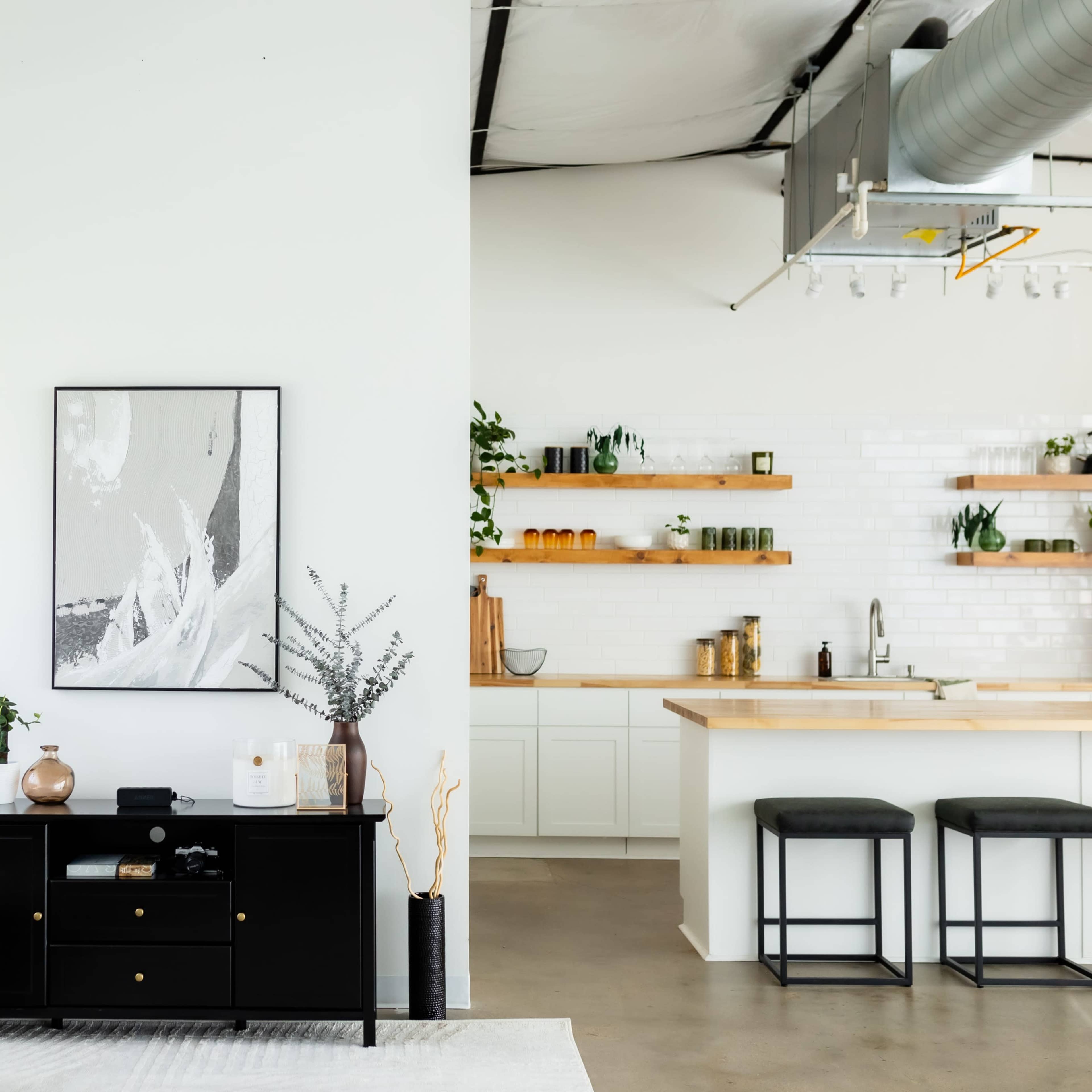 A modern, minimalist living space features a black cabinet, a white wall with a large framed artwork, and a kitchen area with open shelves displaying plants and jars.