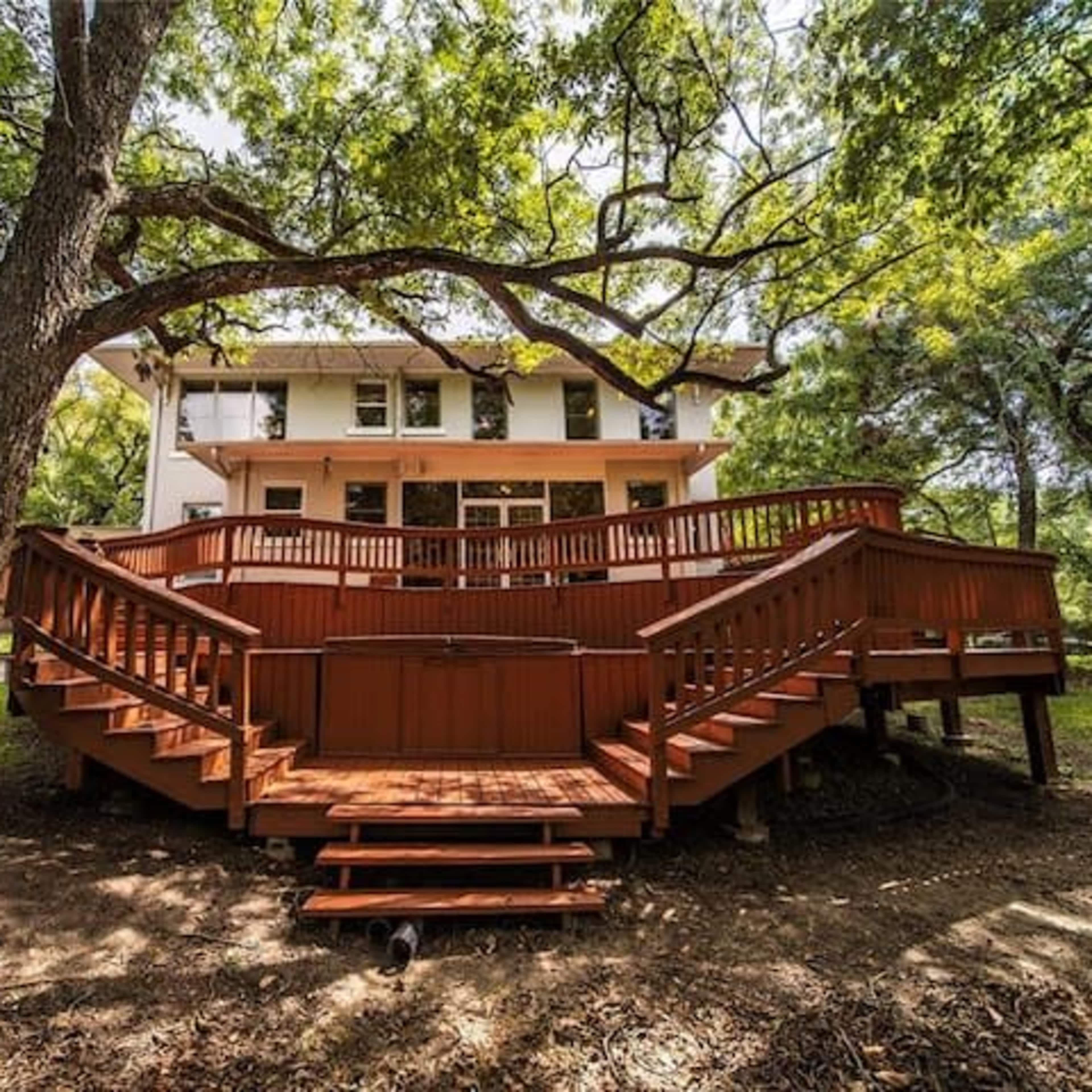 A large wooden deck surrounds the back of a two-story house, set among several trees in a grassy area.