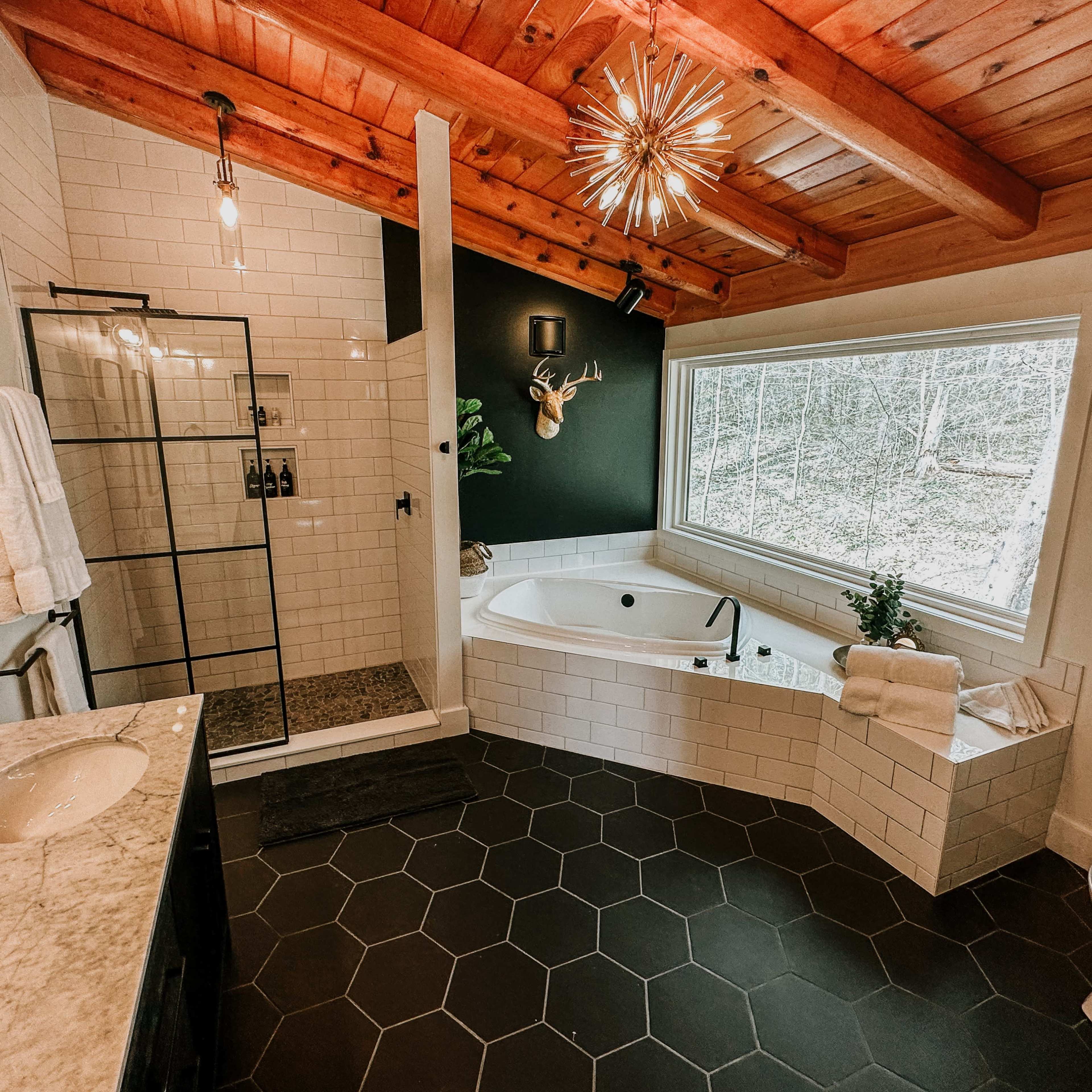A modern bathroom featuring a glass shower, a freestanding bathtub, a double sink vanity with marble countertops, and large windows overlooking a wooded area.