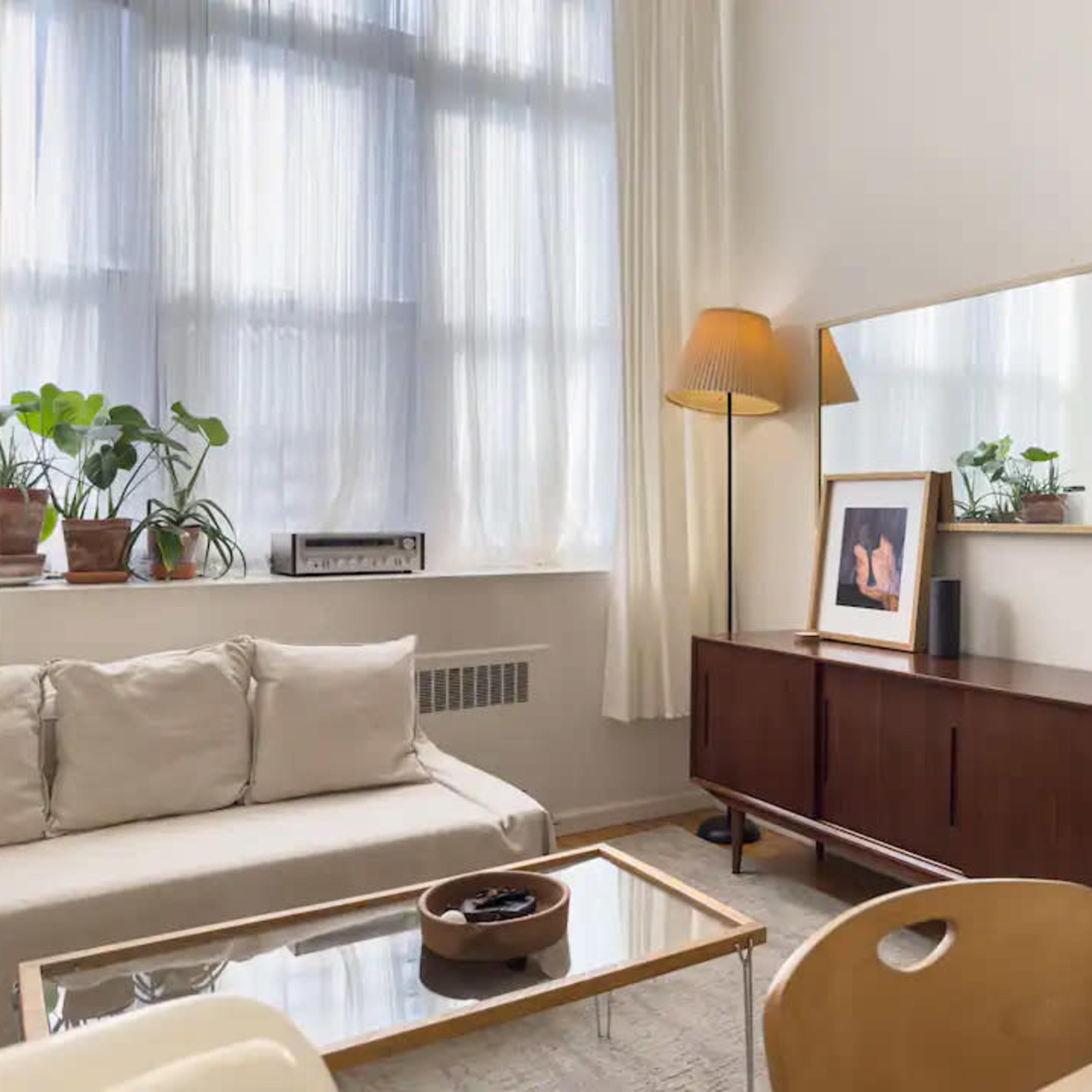 A bright living room featuring a beige sofa, a wooden sideboard with decorative items, a large mirror, and plants near a window with light-colored curtains.
